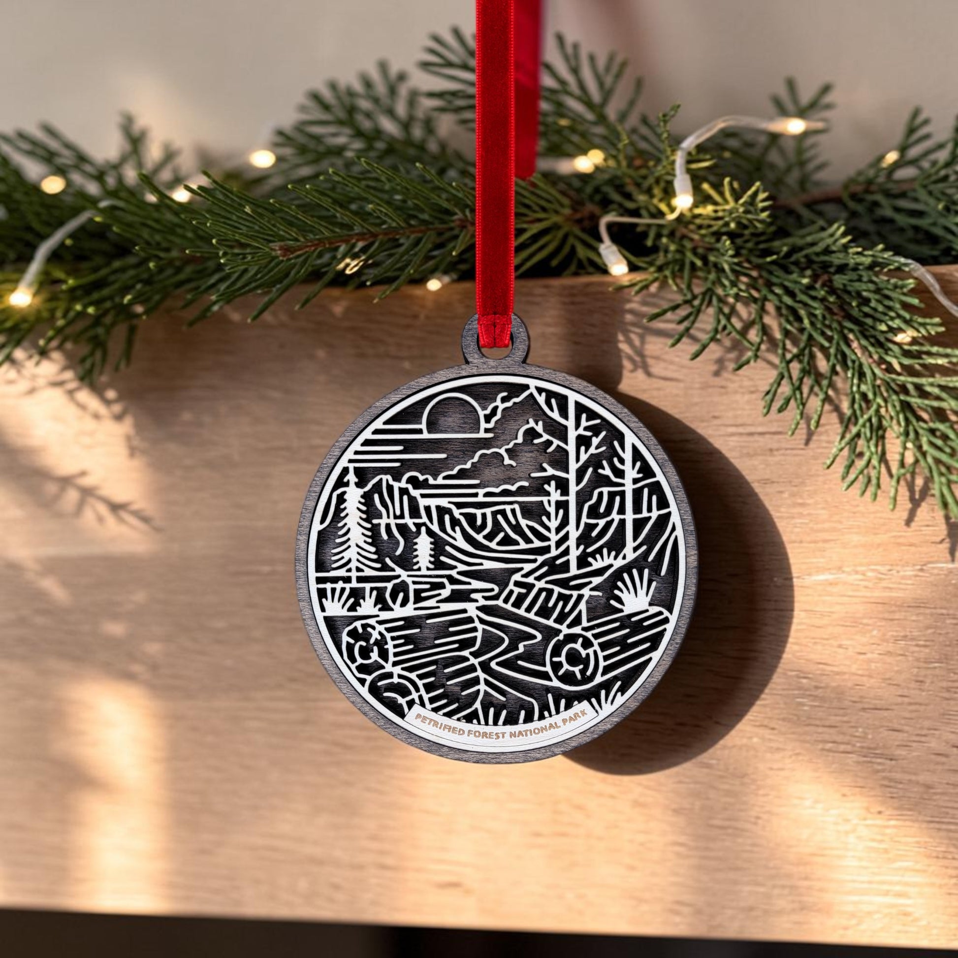 The Petrified Forest National Park Line Art Christmas Ornament by Mirabilia Boutique features an oval wooden design displaying a nature scene with trees, mountains, and a river set against a desert landscape. It hangs from a red ribbon in front of green pine branches and twinkling lights on a wooden surface.