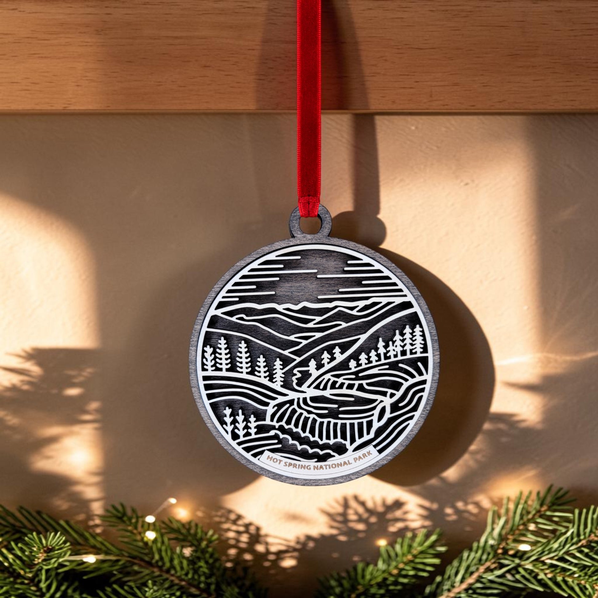 A silver oval Hot Springs National Park Line Art Christmas Ornament by Mirabilia Boutique, featuring an intricately engraved design of mountains, trees, and a road with "Hot Springs National Park" at the bottom, hangs from a red ribbon. Pine branches are visible below, making this ornament a perfect Bathhouse Row keepsake for your collection.