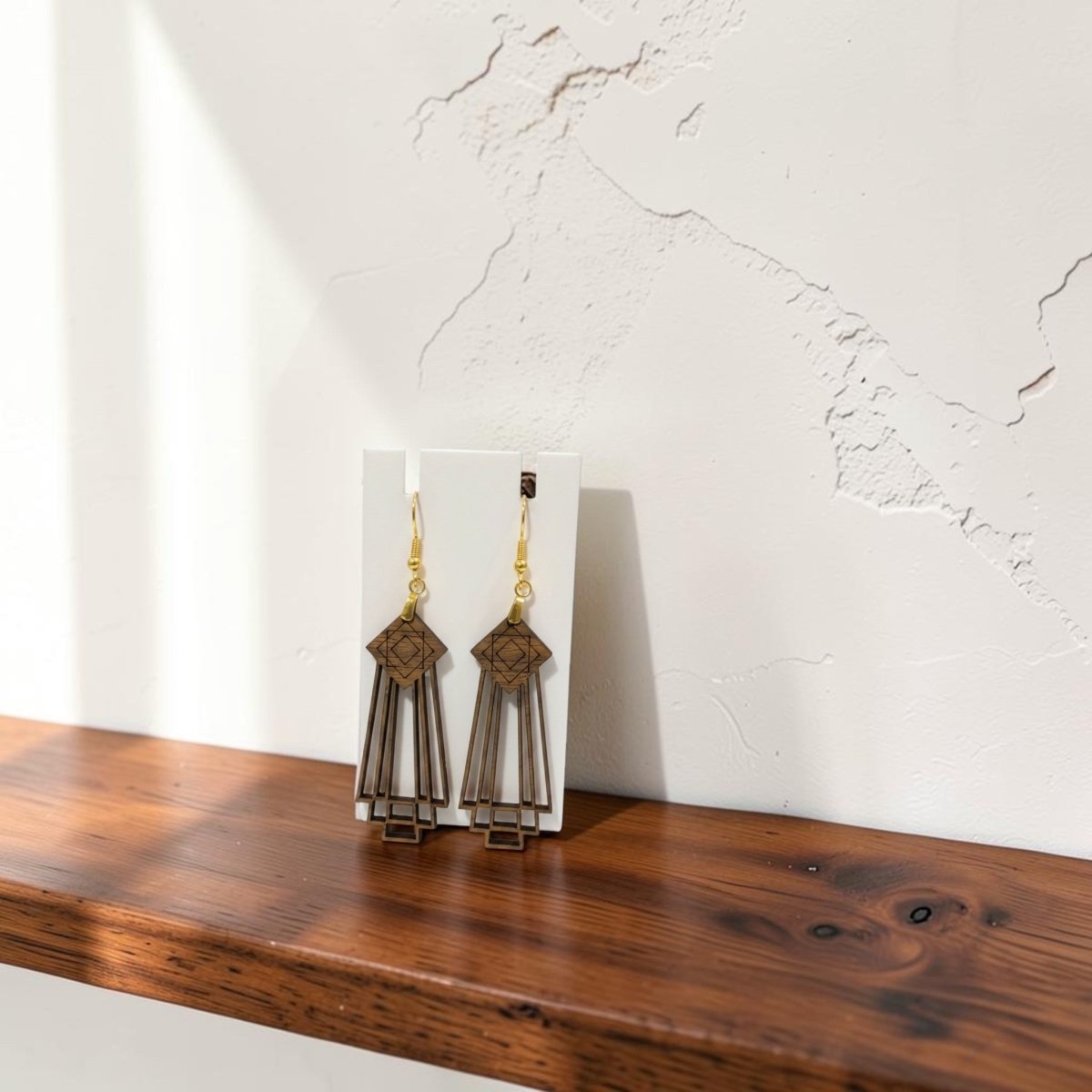 Art Deco Skyscraper Wood Statement Earrings by Mirabilia Boutique with gold hooks are displayed on a white card, resting on a wooden shelf against a white textured wall with subtle cracks. Sunlight casts elegant shadows across the scene, drawing attention to the distinctive Art Deco design and making these Mirabilia Boutique wood statement earrings truly stand out.