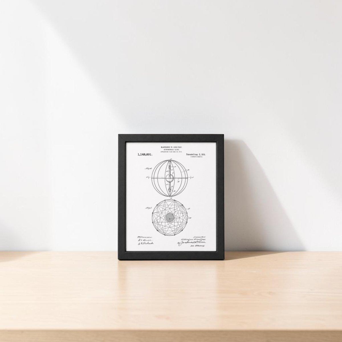The Astronomical Globe Patent Art Print by Mirabilia Boutique is a framed black-and-white vintage-style geometric sphere diagram, displayed on a light wooden table against a white wall with natural lighting and shadows.