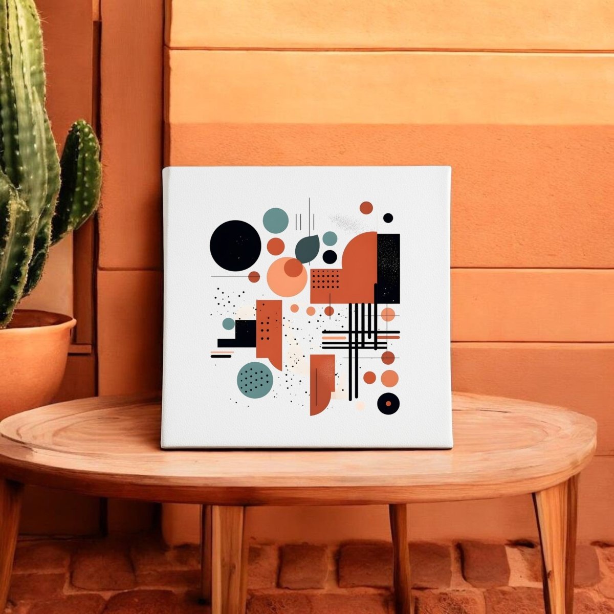 The Atomic Sunrise Mid Century Modern Canvas by Mirabilia Boutique, with retro black, orange, and teal geometry, sits on a round wooden table next to a potted cactus against a terracotta wall.