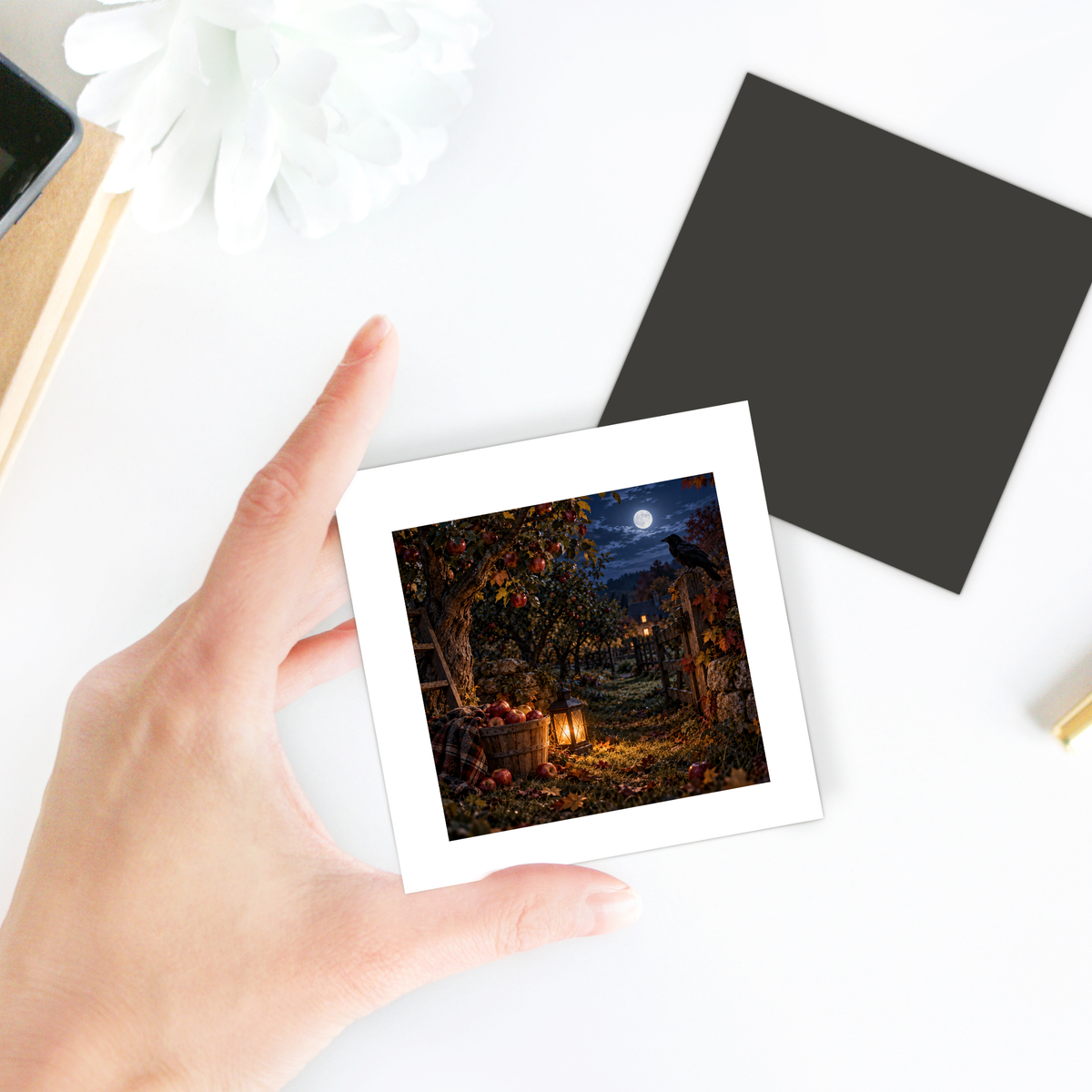 A hand holds a square photo of a lantern-lit apple orchard at night under a full moon—perfect as The Midnight Orchard Magnet | Haunted Harvest from Mirabilia Boutique. Nearby, a blank black square rests on a white surface.