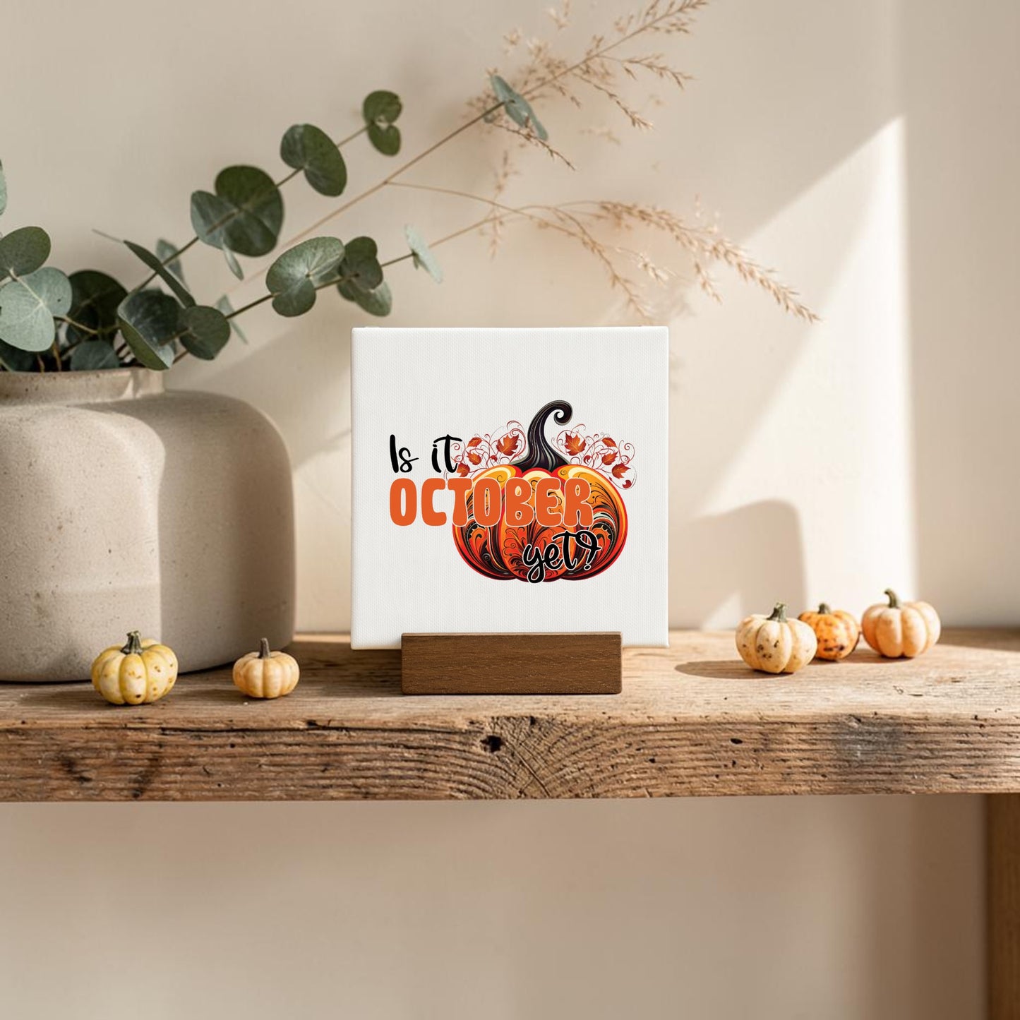 The Is It October Yet? Mini Sign by Mirabilia Boutique features a pumpkin design and playful autumn text, resting on a wooden stand with eucalyptus and mini pumpkins—perfect for charming October decor.