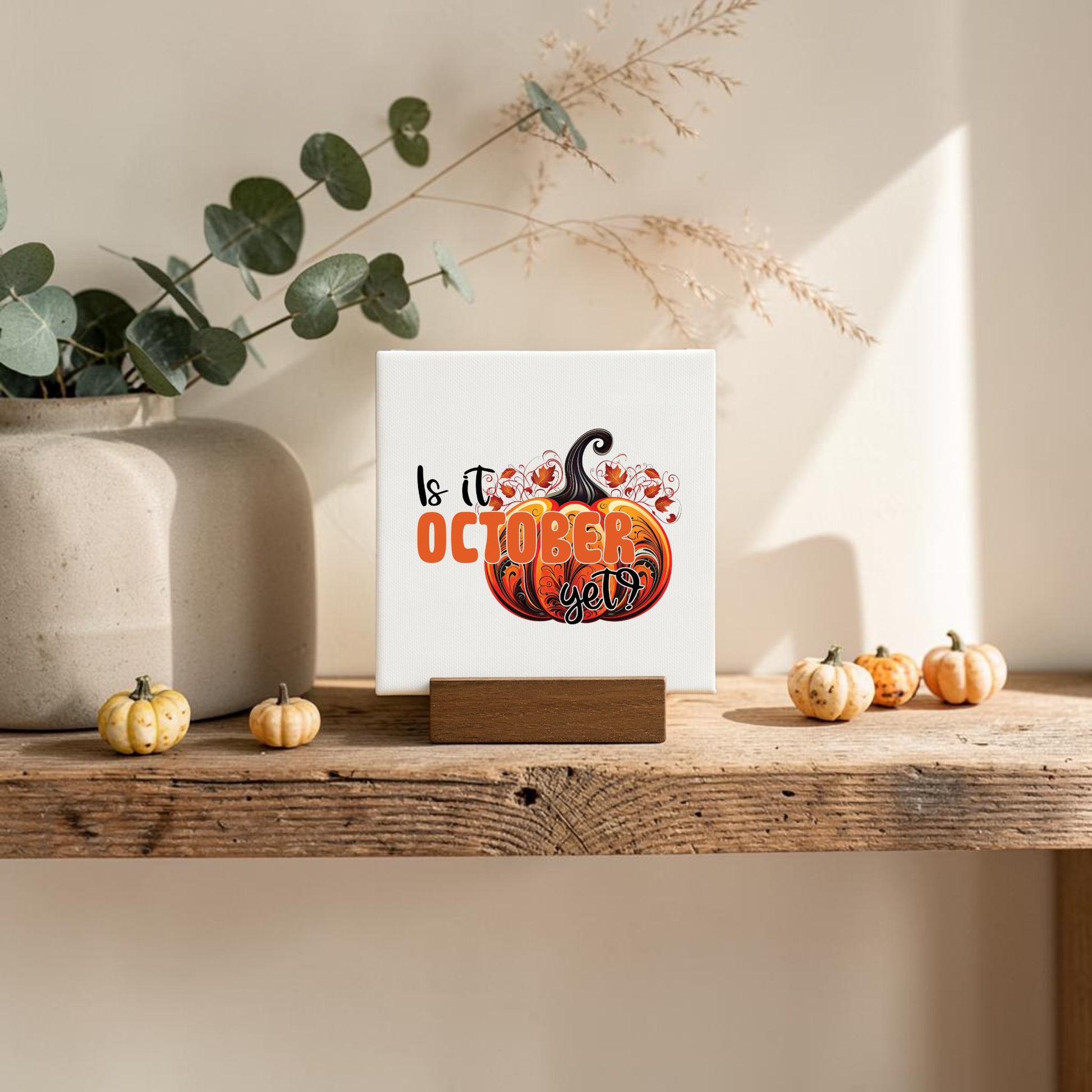 The Is It October Yet? Mini Sign by Mirabilia Boutique features a pumpkin design and playful autumn text, resting on a wooden stand with eucalyptus and mini pumpkins—perfect for charming October decor.