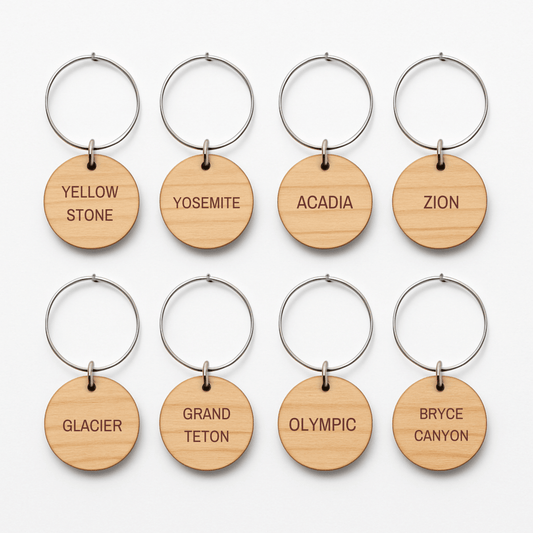Eight round wooden keychains from Mirabilia Boutique's Awe-Inspiring National Parks Wine Charms are arranged in two rows, each engraved with a U.S. national park name—perfect for outdoor entertaining or as unique wine charms.