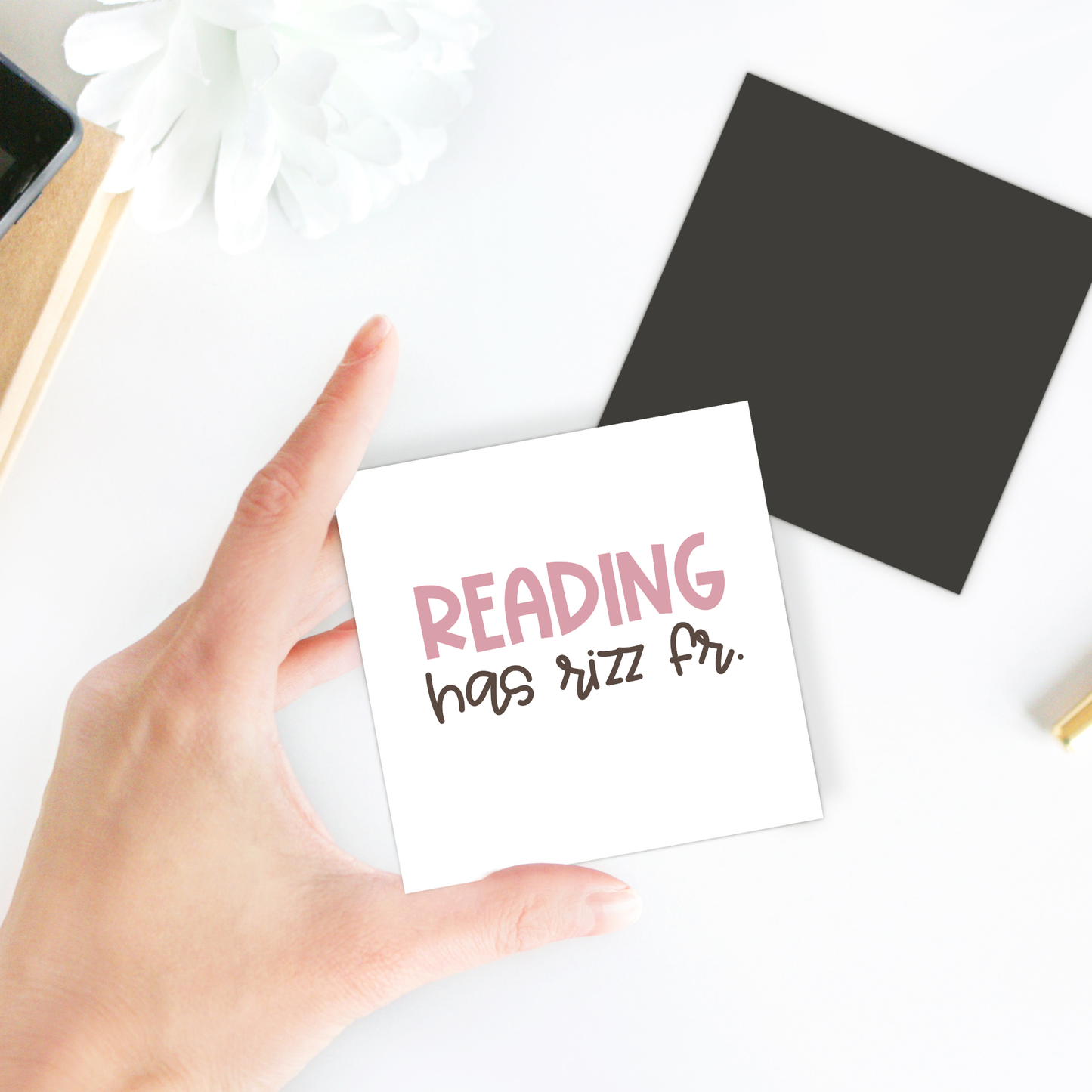 A hand holds the Reading Has Rizz Fr Magnet by Mirabilia Boutique, featuring “READING has rizz fr.” in pink and black. Shown with a white flower, black square, and yellow pencil—great for classroom decor or as a teacher gift.