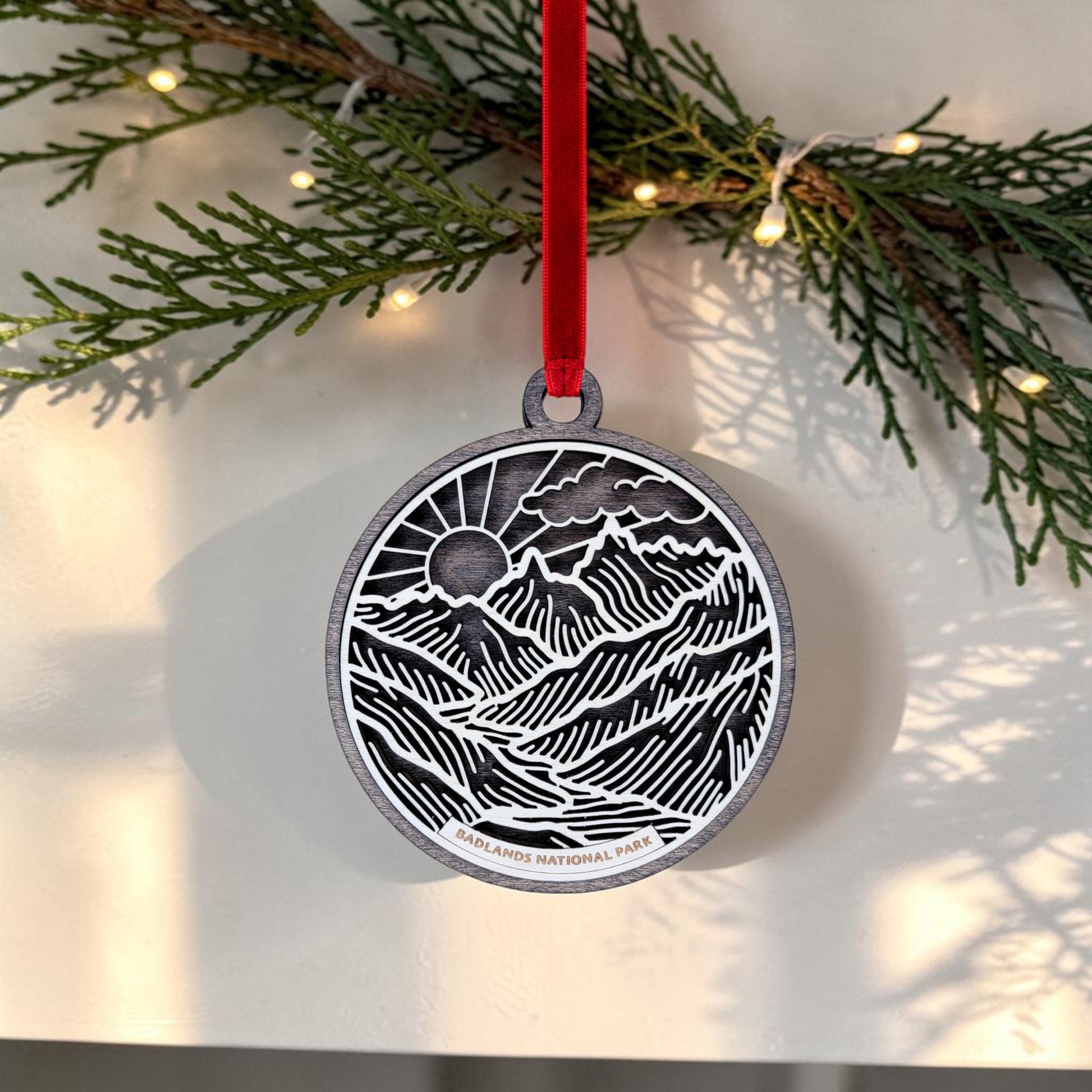 The Badlands National Park Line Art Ornament by Mirabilia Boutique features a round wooden design with a mountain scene and "Badlands National Park" text, hanging from a red ribbon. Pine branches adorned with small white lights provide a festive backdrop, showcasing this distinctive holiday ornament.