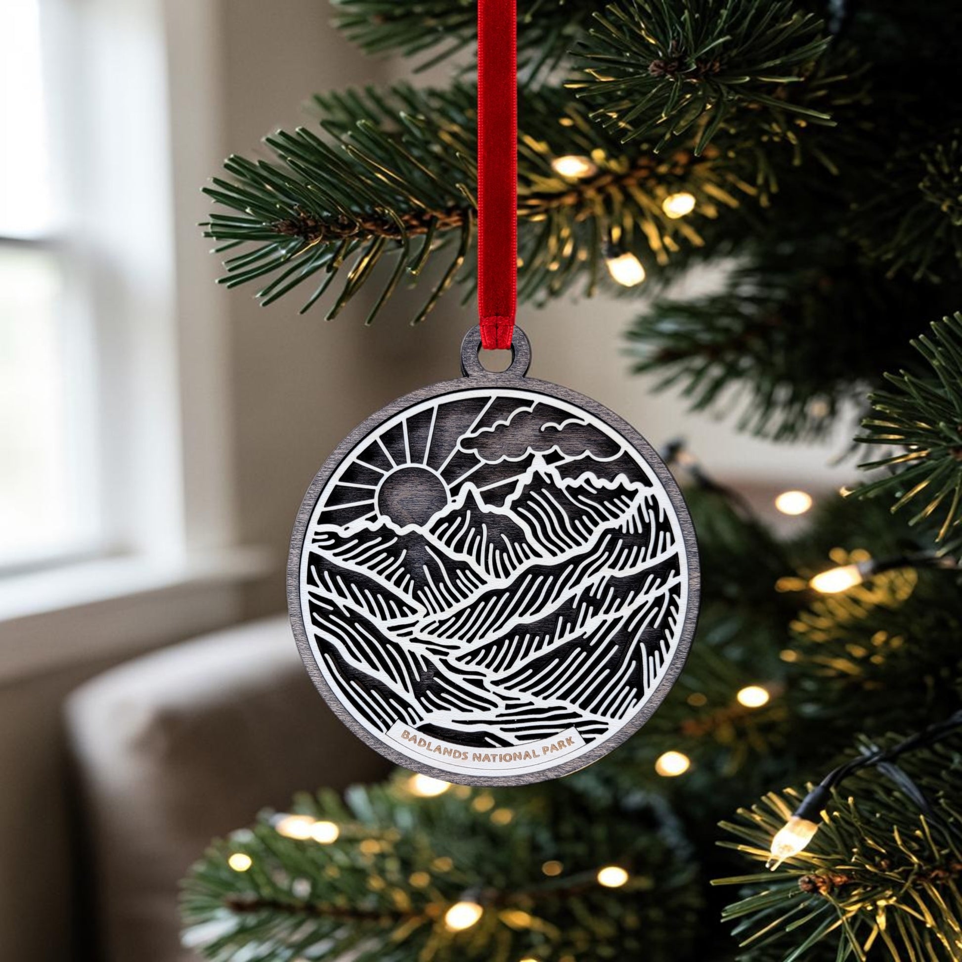 A round metal Badlands National Park Line Art Ornament by Mirabilia Boutique hangs from a red ribbon on a Christmas tree branch with white lights, beautifully evoking the natural landscape of the park.