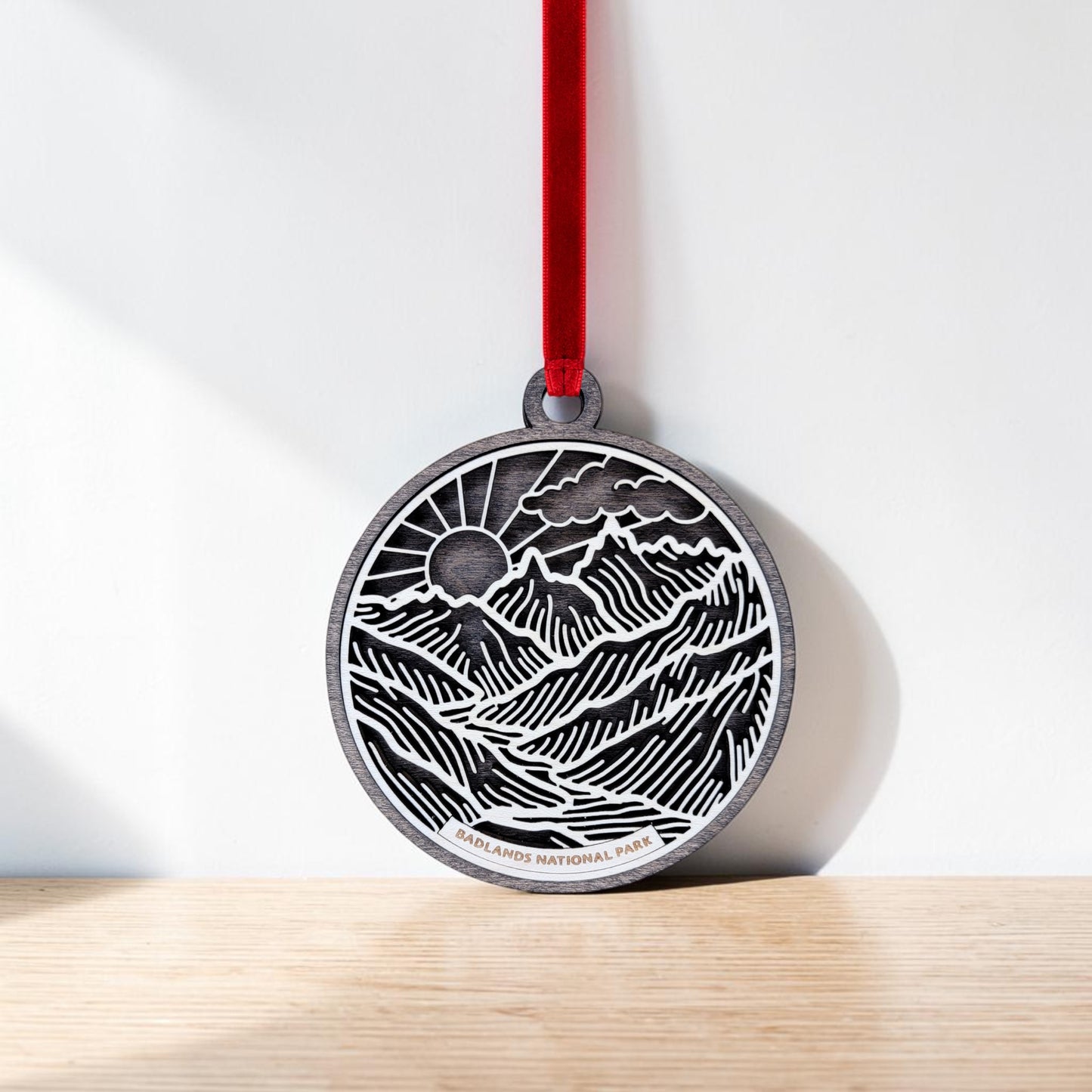 The Badlands National Park Line Art Ornament by Mirabilia Boutique features a round, metallic design adorned with an engraved landscape of mountains, sun, and clouds. Suspended from a red ribbon, this ornament showcases intricate line art that beautifully represents the essence of Badlands National Park. "Badlands National Park" is elegantly etched at the bottom of the piece. Displayed against a light-colored wall on a wooden surface, this ornament highlights both craftsmanship and natural beauty.