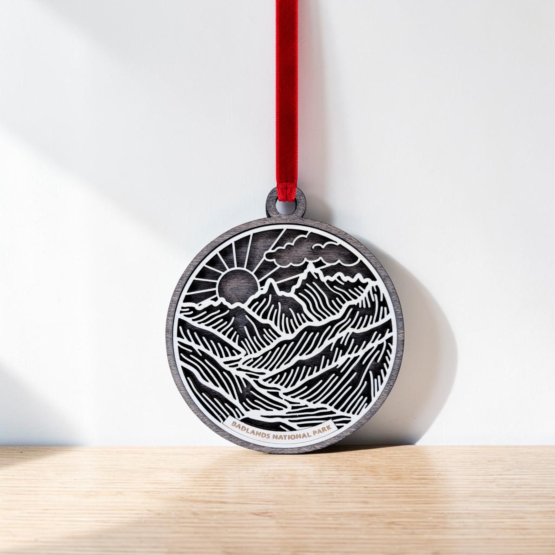 The Badlands National Park Line Art Ornament by Mirabilia Boutique features a round, metallic design adorned with an engraved landscape of mountains, sun, and clouds. Suspended from a red ribbon, this ornament showcases intricate line art that beautifully represents the essence of Badlands National Park. "Badlands National Park" is elegantly etched at the bottom of the piece. Displayed against a light-colored wall on a wooden surface, this ornament highlights both craftsmanship and natural beauty.