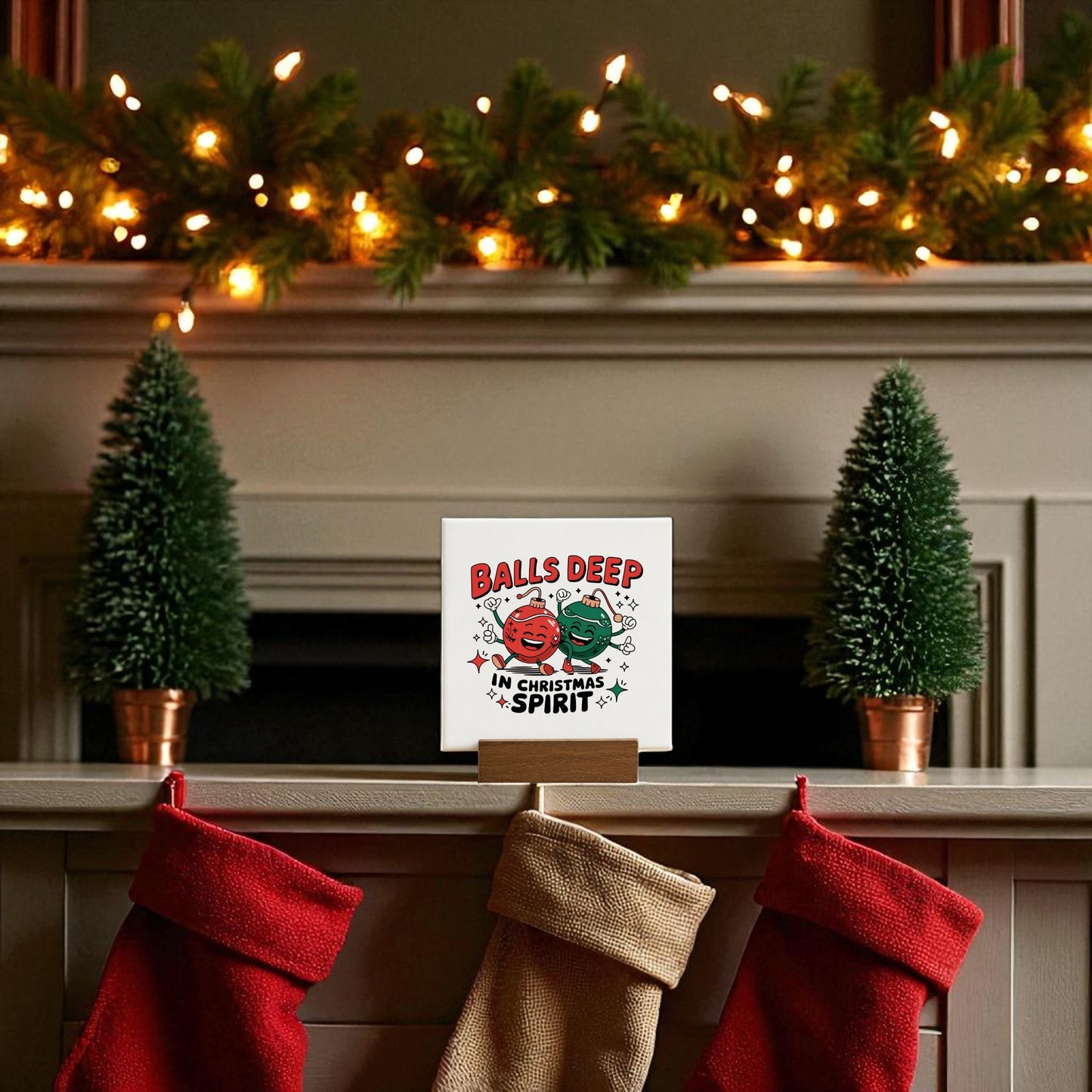 A festive mantel with two mini Christmas trees, three stockings, twinkling lights, garland, and the Mirabilia Boutique "Balls Deep in Christmas Spirit" Funny Mini Holiday Sign for playful adult holiday decor.