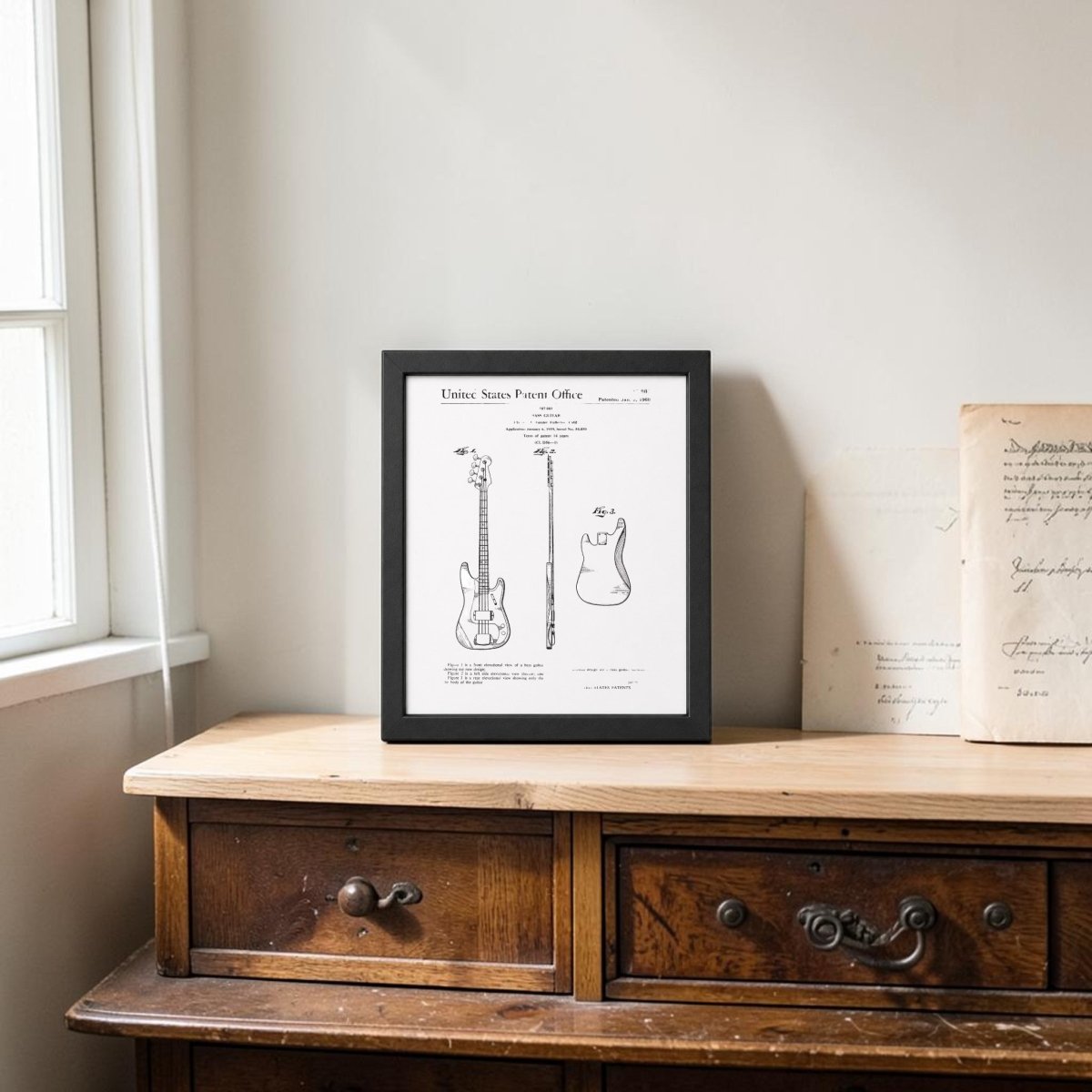The Mirabilia Boutique Bass Guitar Patent Print on Metal (8x10 Wall Art) adds vintage charm to a light-walled room, displayed on a wooden dresser beside old papers with sunlight streaming in. Perfect music decor for any stylish space.