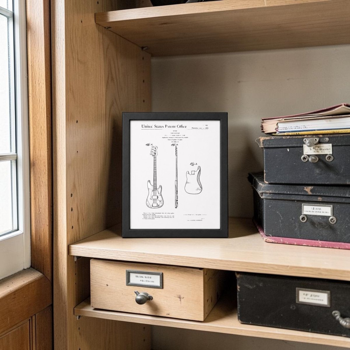 The Mirabilia Boutique Bass Guitar Patent Print on Metal (8x10) rests on a wooden shelf near black boxes, folders, and a window, with natural light highlighting this unique piece of music wall art.