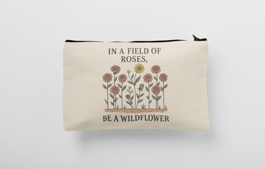 The Be a Wildflower Zipper Pouch by Mirabilia Boutique features pink roses, a yellow wildflower, and the quote "In a field of roses, be a wildflower." This versatile canvas bag is perfect as an inspirational makeup bag or pencil case.