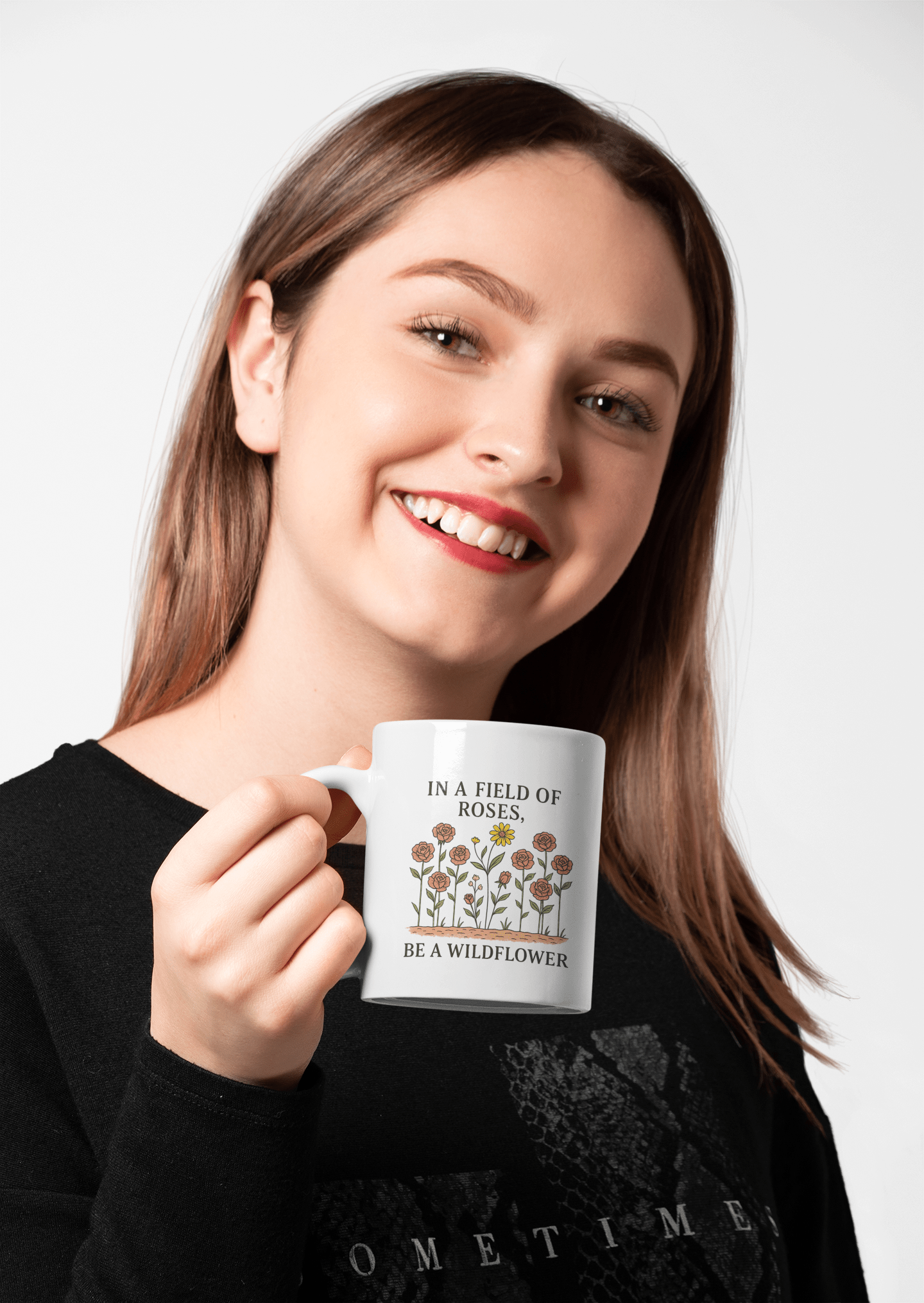 A smiling woman with long brown hair holds the "Be a Wildflower Mug | Inspirational Ceramic Coffee Cup" from Mirabilia Boutique— a white mug with floral design and the phrase, "In a field of roses, be a wildflower." She wears black, standing against a light background.