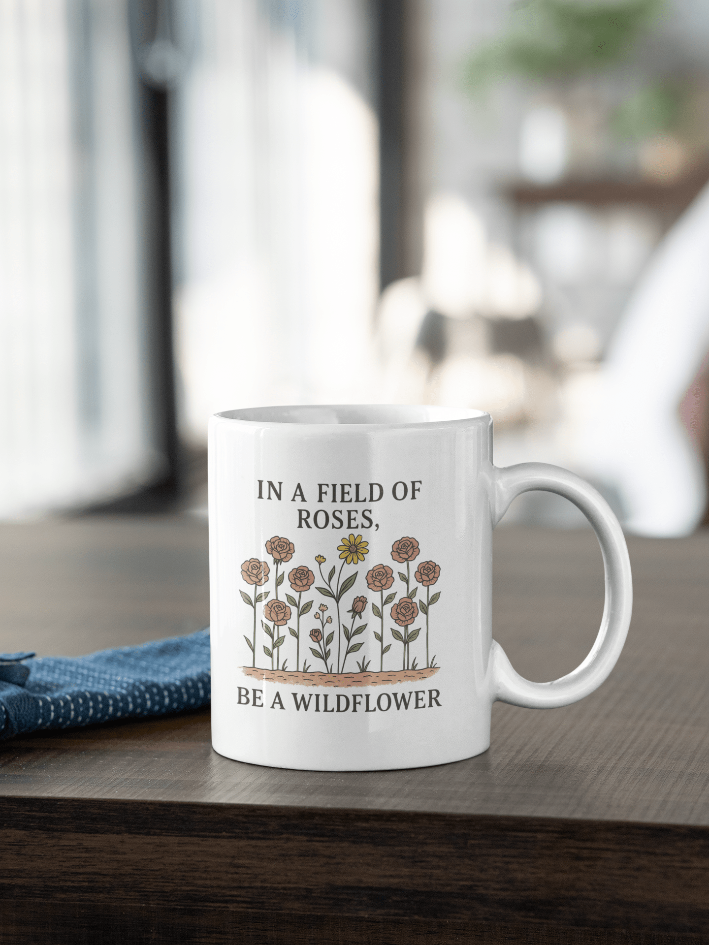The Be a Wildflower Mug by Mirabilia Boutique is a white ceramic coffee cup with a motivational floral design and the quote "In a field of roses, be a wildflower," perfect for cozy indoor moments.