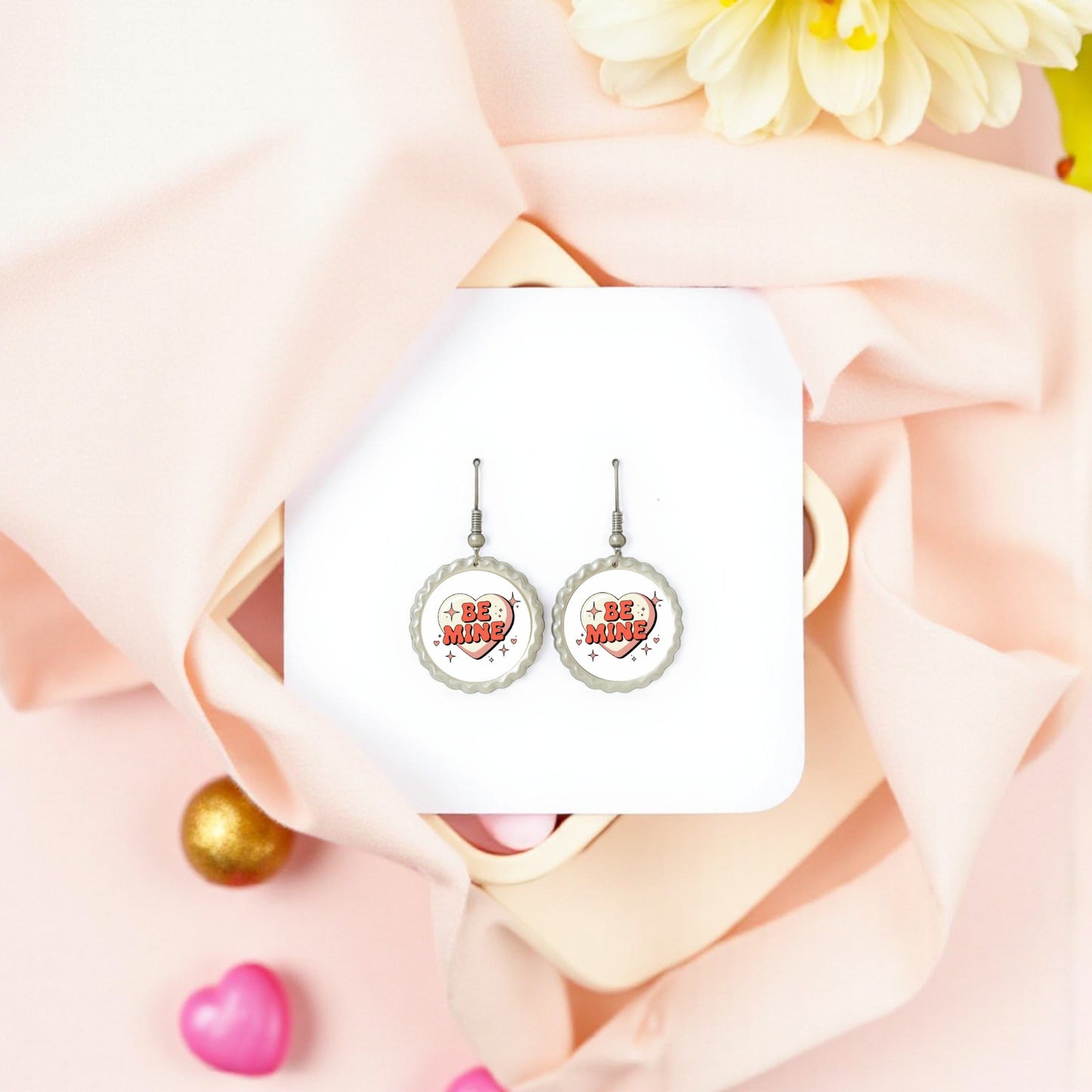 Be Mine Heart Earrings by Mirabilia Boutique, a cute Valentine jewelry gift, are shown on a white square with pink fabric, a cream flower, and heart-shaped decor—perfect retro-style earrings for Valentine’s Day.