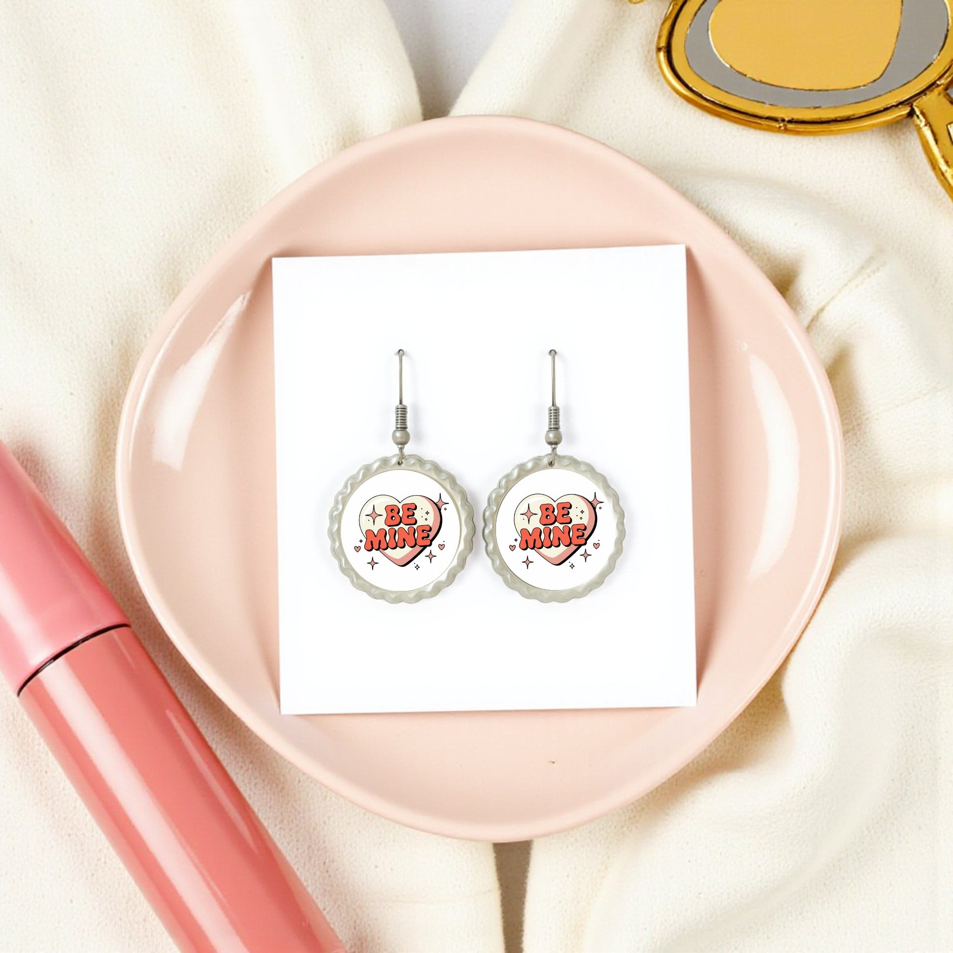 Be Mine Heart Earrings by Mirabilia Boutique are shown on a white card atop a pink plate, with a pink tube and heart mirror, creating a charming retro vibe. This cute Valentine jewelry gift is styled on a cream fabric background.