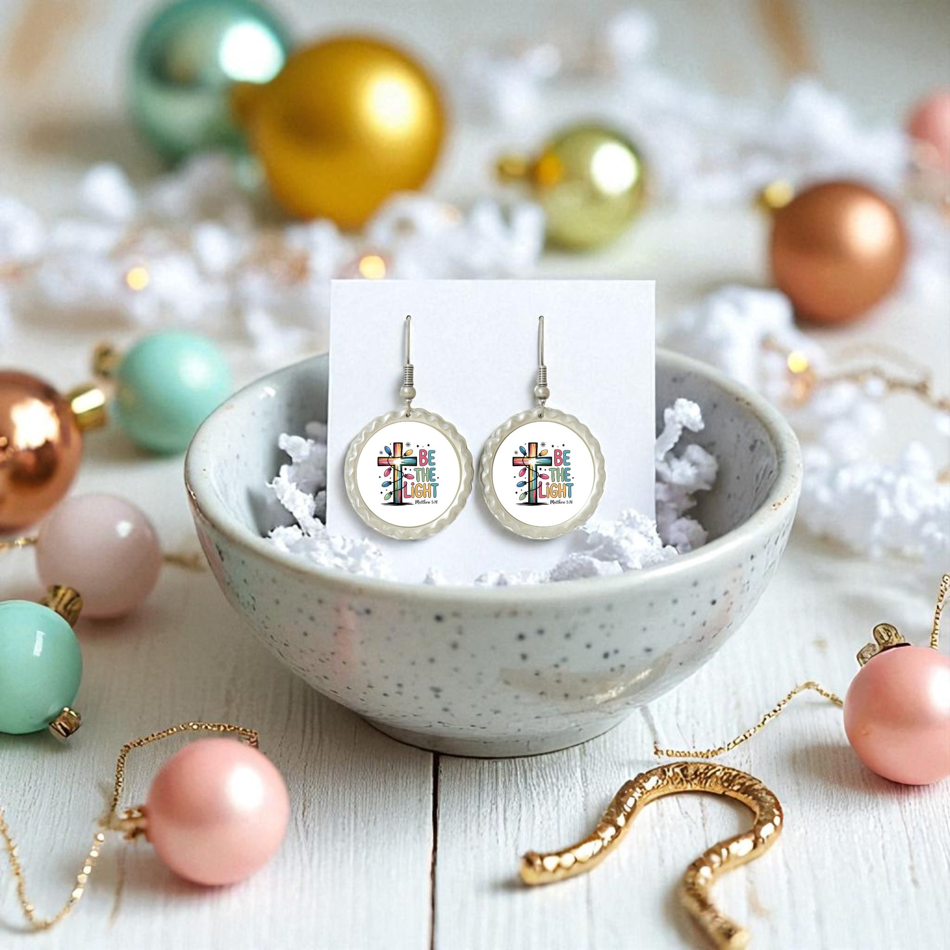 The Be the Light Earrings by Mirabilia Boutique feature a colorful cross and “Be the Light,” displayed on a white card in a speckled bowl amid pastel ornaments and gold necklaces. A faith-filled Christian gift inspired by Matthew 5:14.