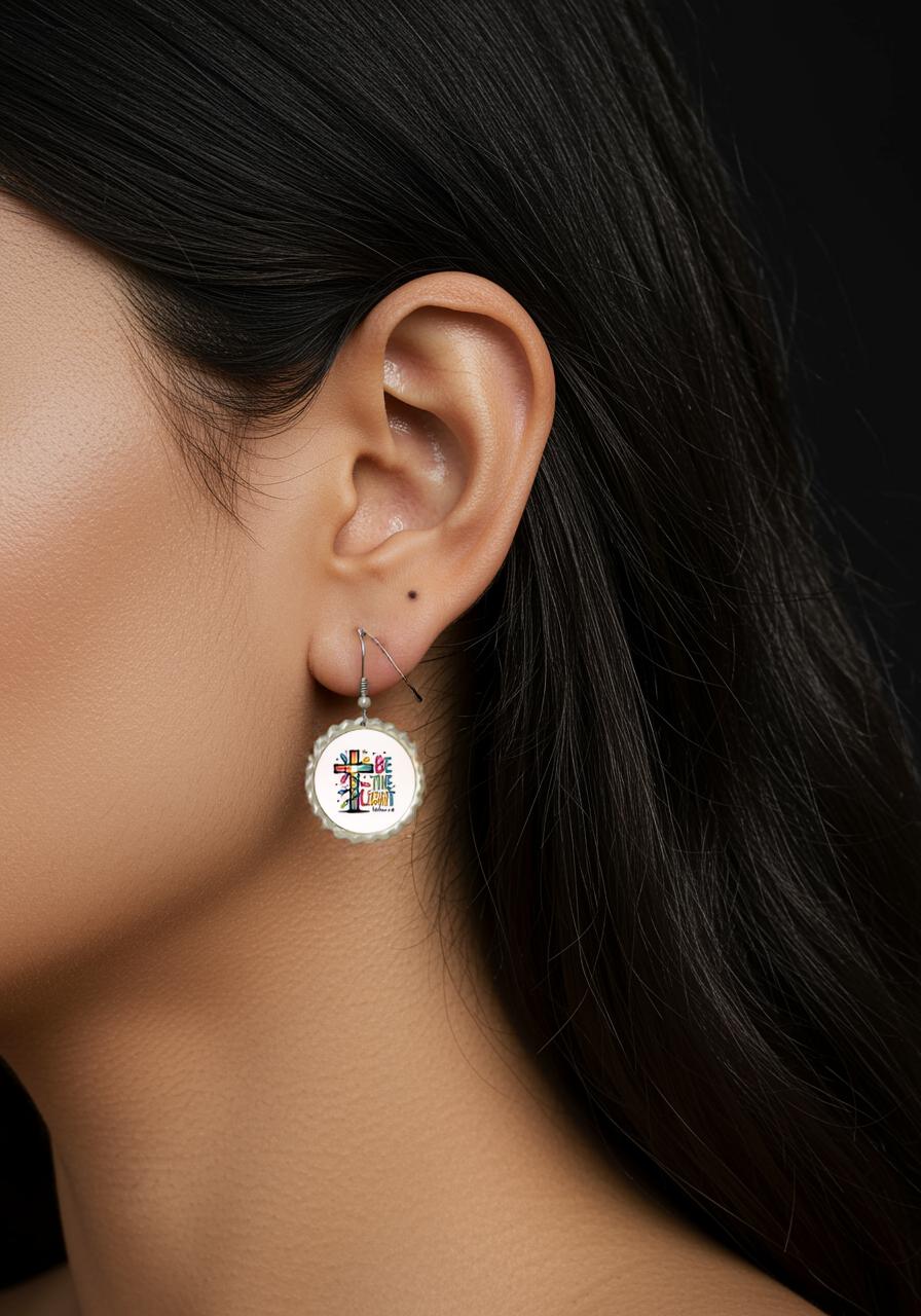Close-up of a woman’s ear with long dark hair, wearing Mirabilia Boutique’s “Be the Light Earrings”—a round, white piece with a colorful abstract design inspired by Matthew 5:14. The faith-filled Christian gift stands out against the black background.
