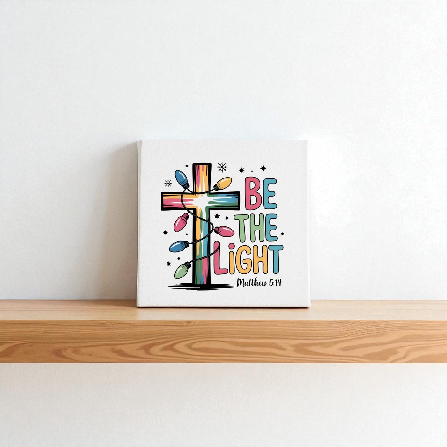 The Be the Light canvas by Mirabilia Boutique features Matthew 5:14 with colorful Christmas décor, string lights, stars, and playful lettering—a perfect faith-inspired holiday accent for your shelf.