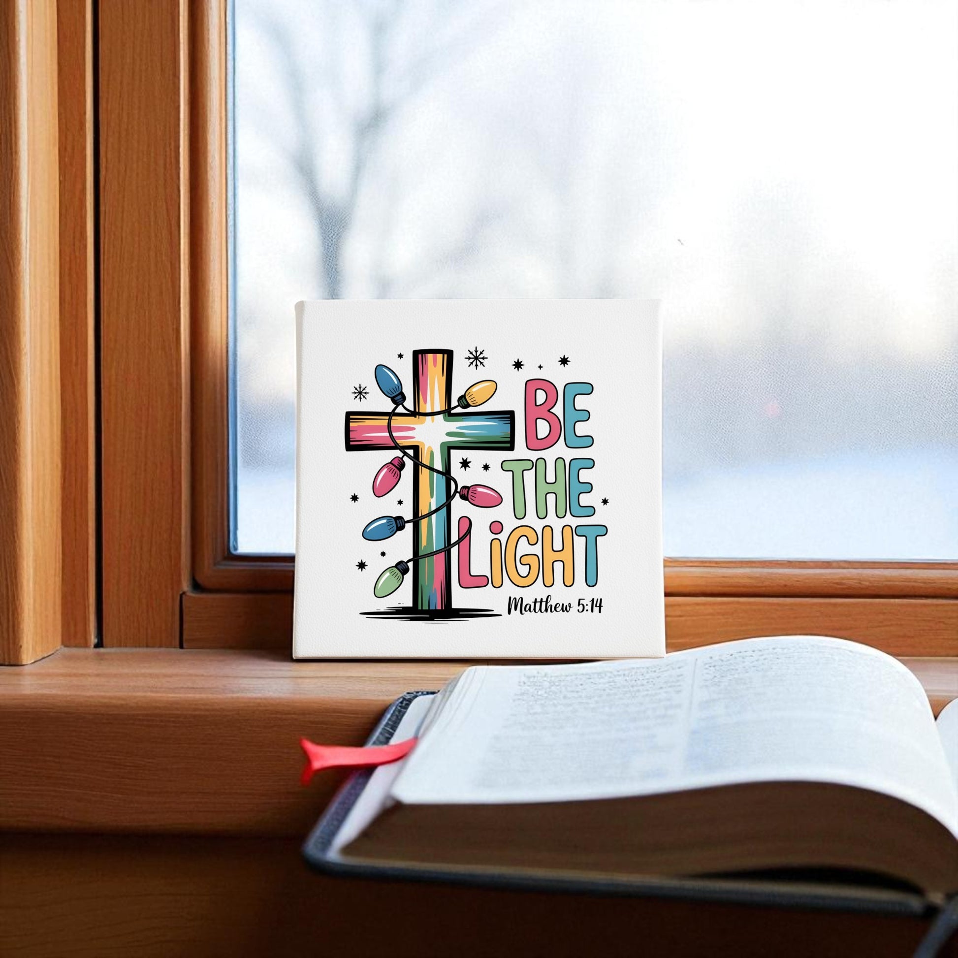 The "Be the Light | Faith-Inspired Christmas Decor | Matthew 5:14" sign by Mirabilia Boutique sits on a windowsill beside an open Bible, with snowy trees outside, creating a festive, faith-inspired holiday scene.