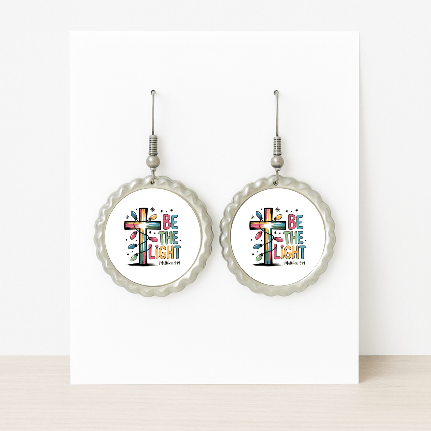 A pair of "Be the Light" earrings by Mirabilia Boutique featuring a colorful cross and Matthew 5:14—Christian gifts displayed on a card with scalloped silver edges, perfect for faith-inspired giving.