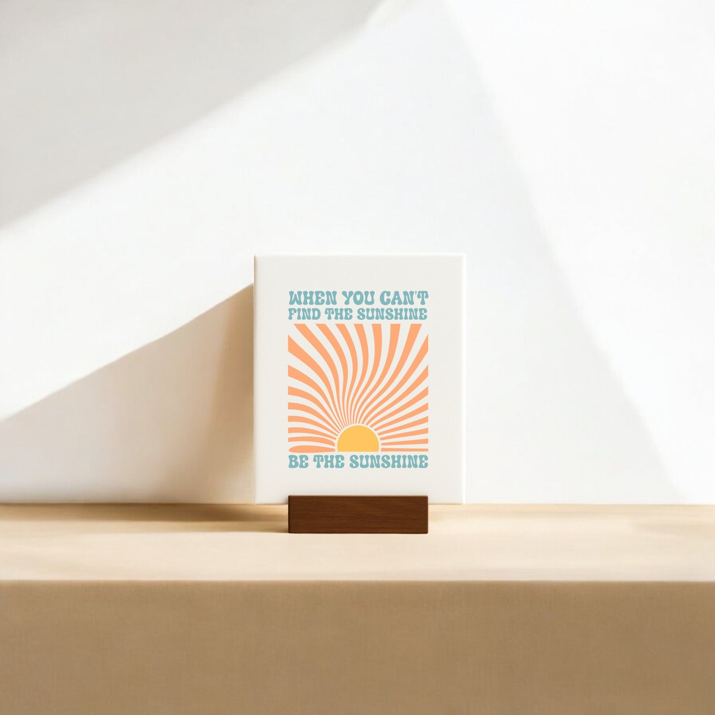 The Be the Sunshine Mini Canvas – Retro Sunburst Sign by Mirabilia Boutique features an orange sunburst and “WHEN YOU CAN'T FIND THE SUNSHINE, BE THE SUNSHINE” quote on a wooden stand for inspiring, neutral-toned decor.