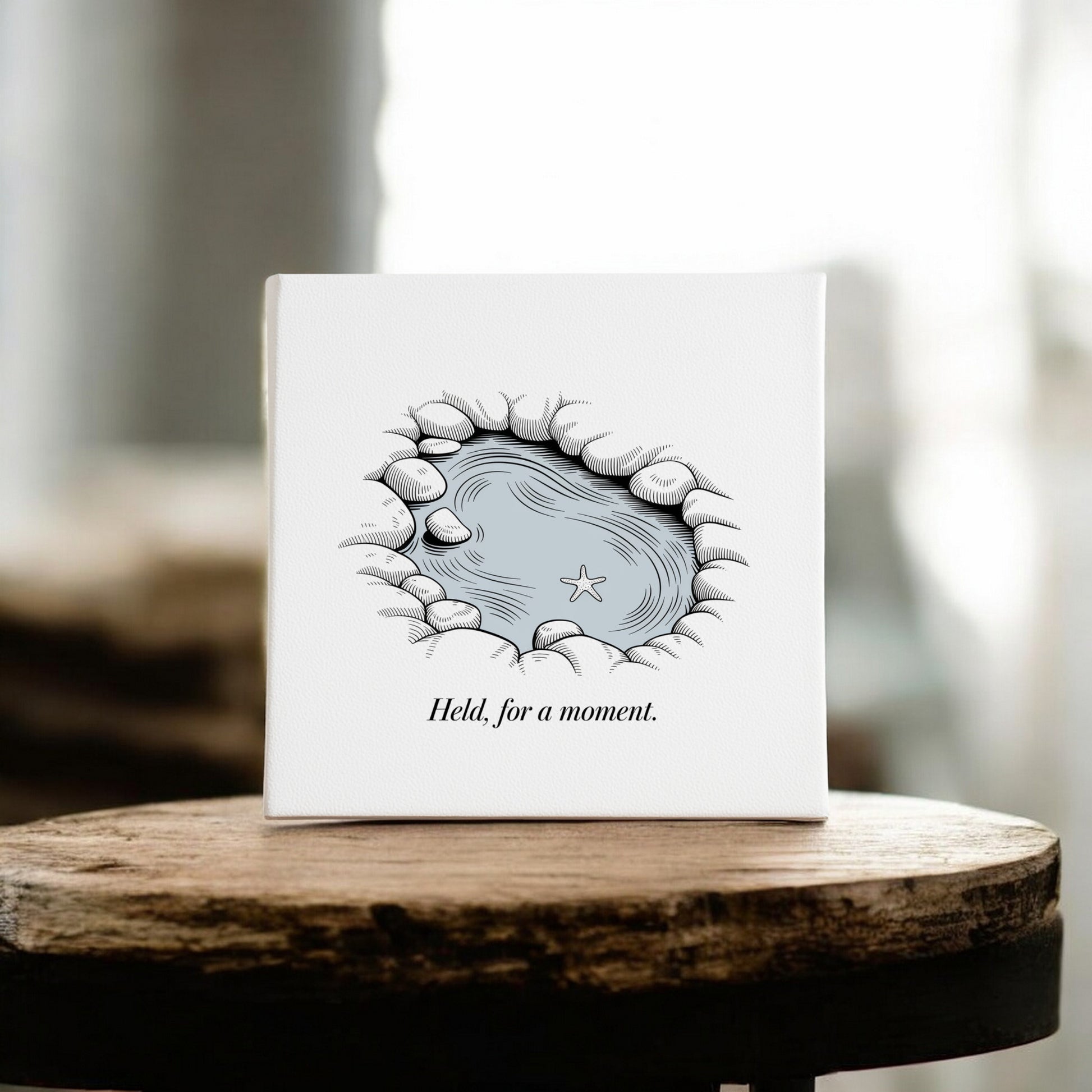 The “Held, For a Moment – 8x8 Coastal Canvas Wall Art” by Mirabilia Boutique features a rocky tidepool scene with a starfish, set on a wooden stool. The serene artwork displays the phrase, “Held, for a moment.”.