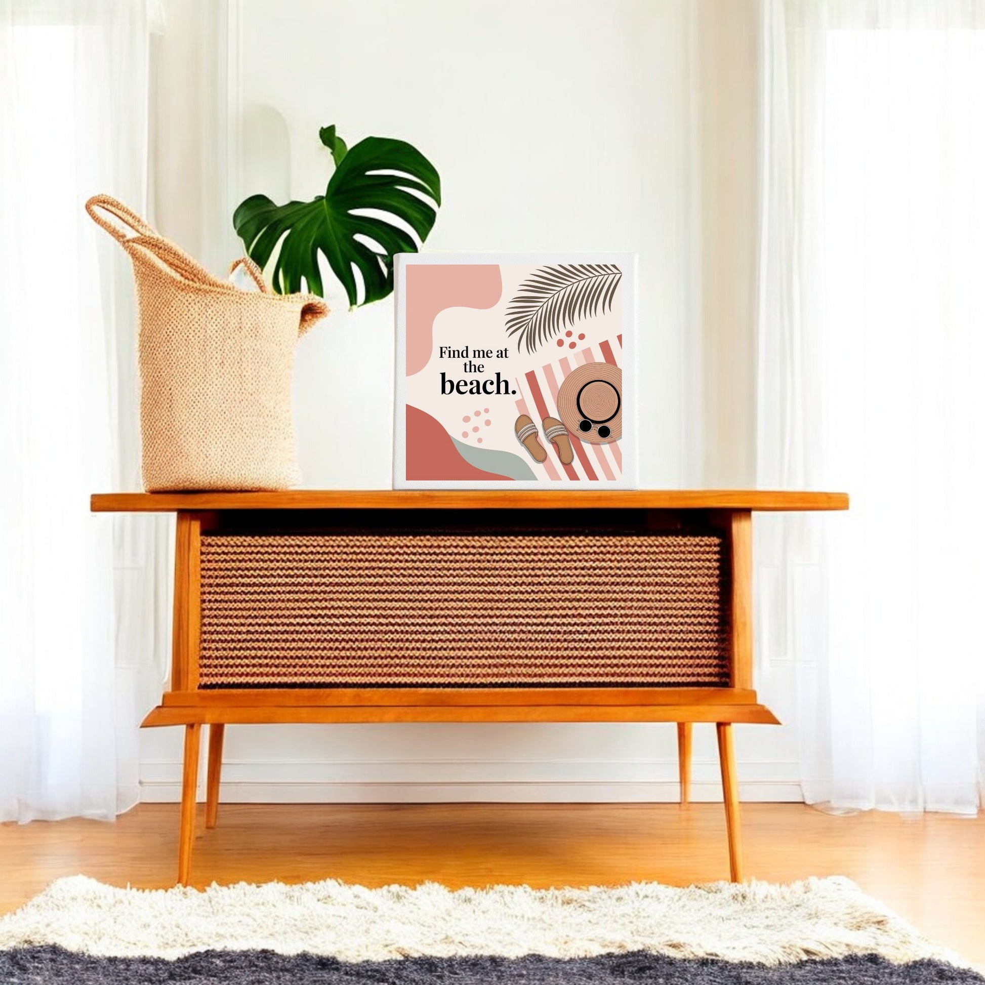A mid-century wooden console stands in a bright room with sheer curtains, topped with a woven basket, monstera leaf, and Mirabilia Boutique’s “Find Me at the Beach Canvas – Coastal Summer Vibes” wall art.