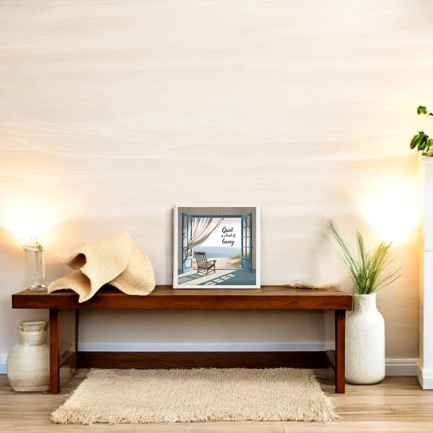 A minimalist room features a wooden bench, a woven hat, and a plant. The Mirabilia Boutique “Quiet Is a Kind of Luxury Canvas – Peaceful Summer Wall Art” hangs above, softly lit by twin lamps for an atmosphere of serene elegance.