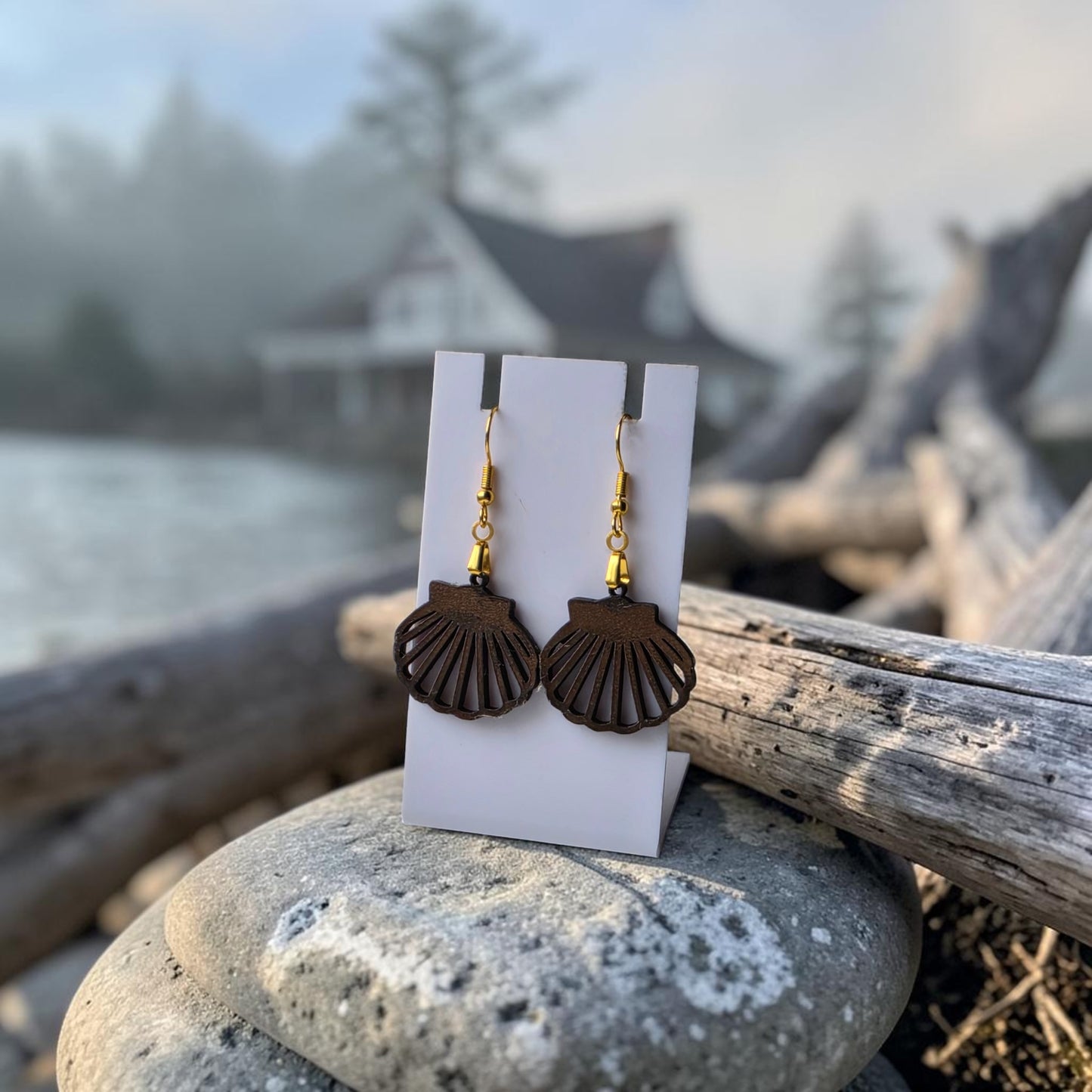 Scallop Shell Earrings by Mirabilia Boutique, crafted from lasercut walnut wood with gold hooks, are displayed on a white card among smooth rocks—perfect coastal jewelry for a seaside-inspired style.