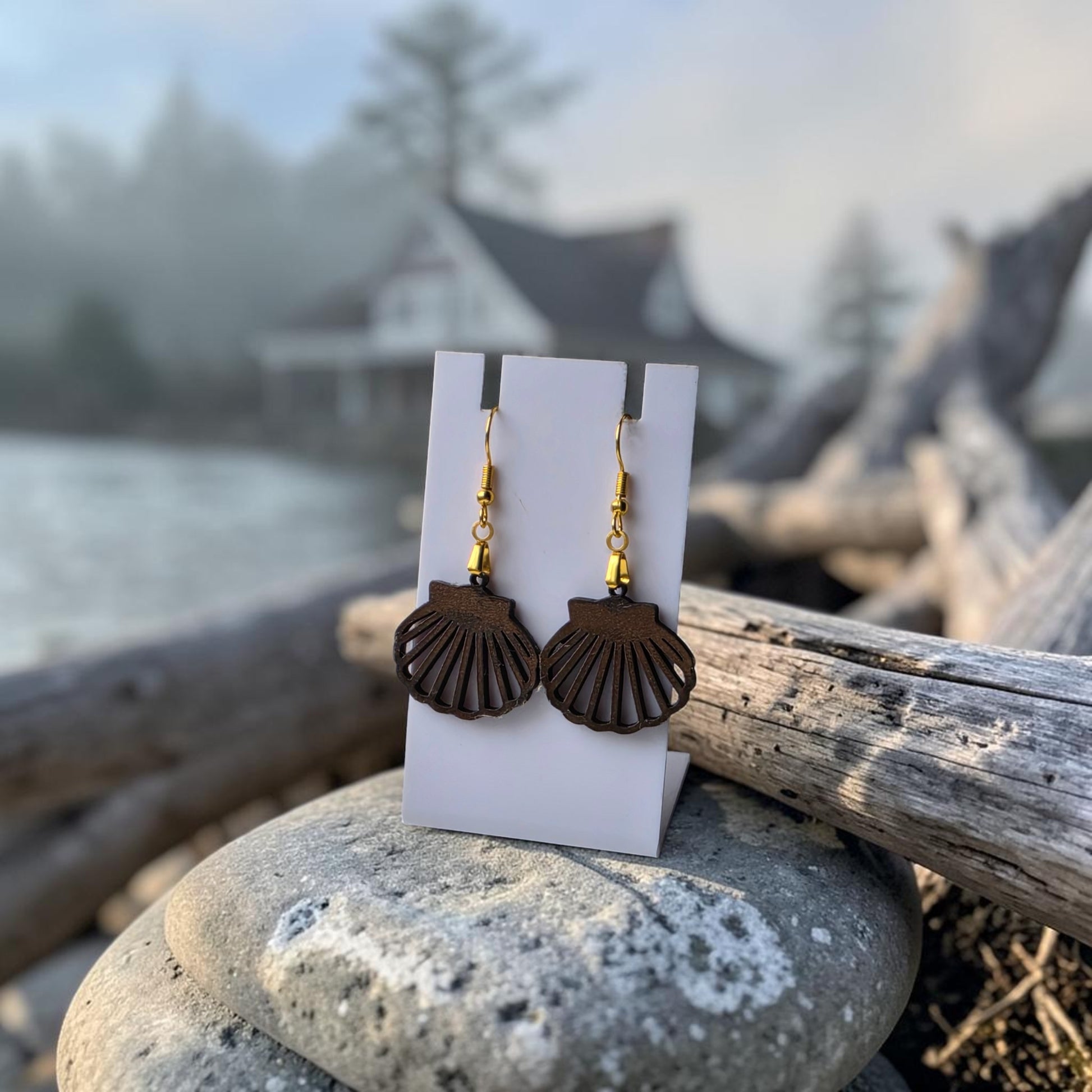 Scallop Shell Earrings by Mirabilia Boutique, crafted from lasercut walnut wood with gold hooks, are displayed on a white card among smooth rocks—perfect coastal jewelry for a seaside-inspired style.