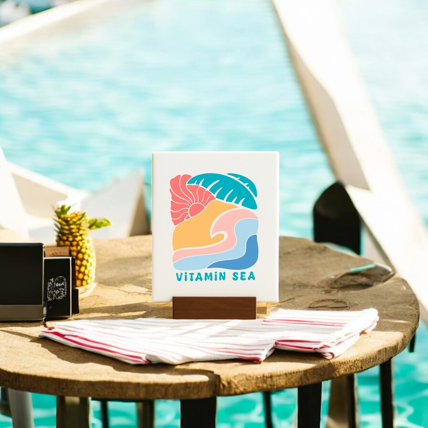 Mirabilia Boutique’s Vitamin Sea Mini Canvas Sign (4x6) adds a pop of coastal decor to a round wooden poolside table, styled with a pineapple, striped cloth, and small chalkboard as sunlight sparkles on the water.
