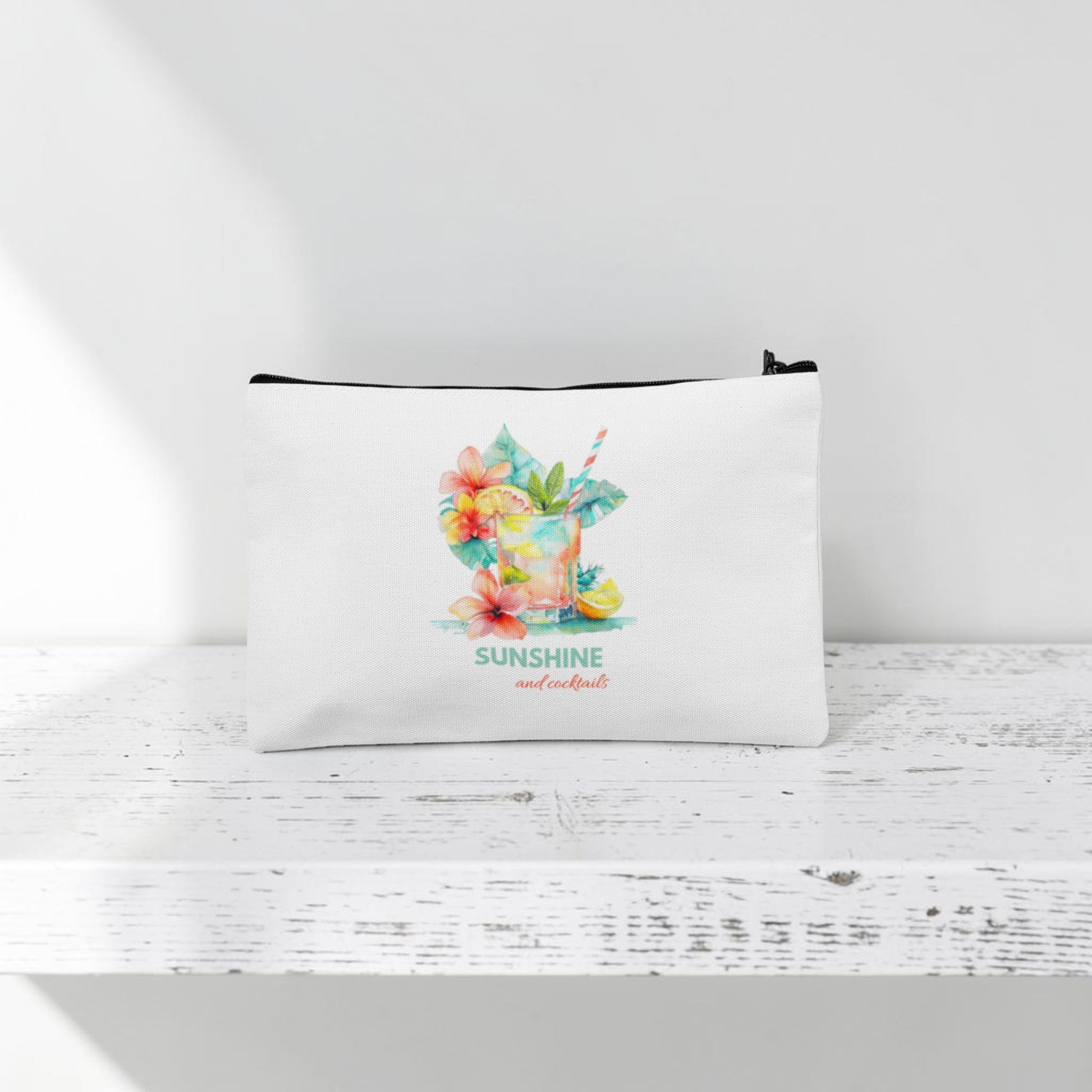 The Mirabilia Boutique Sunshine and Cocktails Makeup Bag—a white zippered travel pouch featuring a vibrant tropical drink, colorful flowers, and the phrase "SUNSHINE and cocktails" in decorative text—rests on a white wooden surface with a white background.