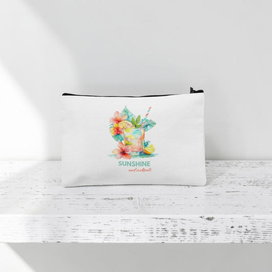 The Mirabilia Boutique Sunshine and Cocktails Makeup Bag—a white zippered travel pouch featuring a vibrant tropical drink, colorful flowers, and the phrase "SUNSHINE and cocktails" in decorative text—rests on a white wooden surface with a white background.