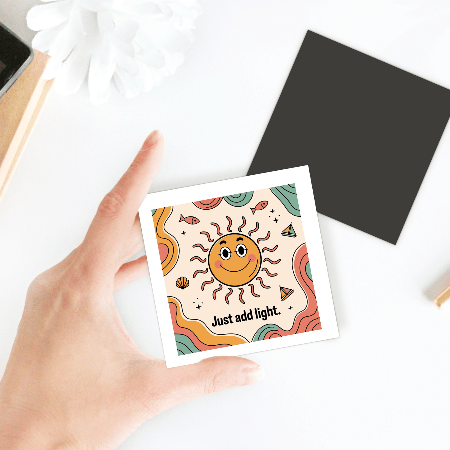 A hand holds Mirabilia Boutique’s “Just Add Light Magnet – Retro Sun Fridge Art,” featuring a smiling sun and vibrant colors. The playful design, with a wavy background and white flower, brings cheerful energy to any space.