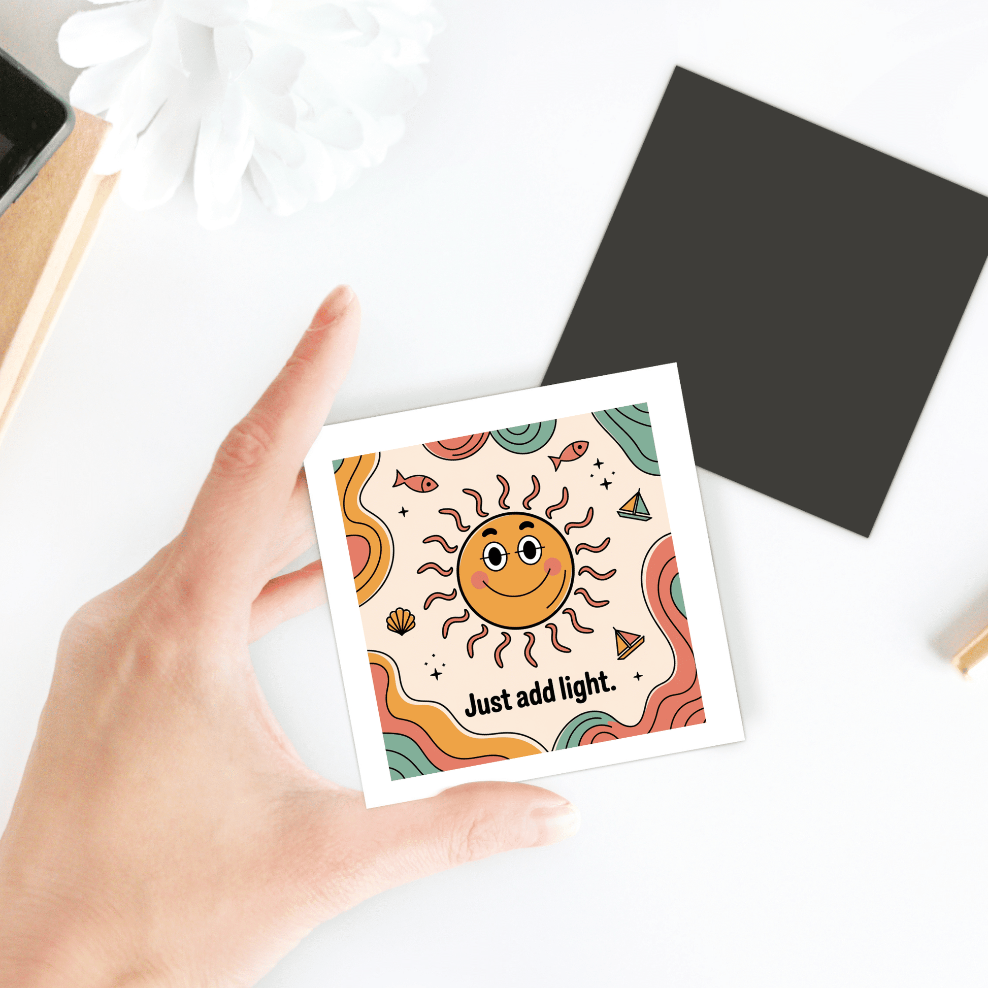 A hand holds Mirabilia Boutique’s “Just Add Light Magnet – Retro Sun Fridge Art,” featuring a smiling sun and vibrant colors. The playful design, with a wavy background and white flower, brings cheerful energy to any space.