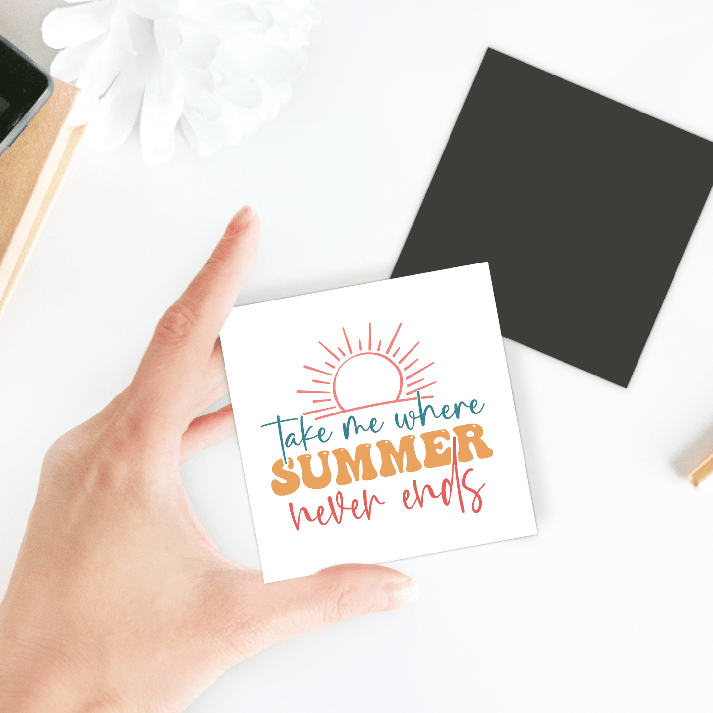 A hand holds the "Take Me Where Summer Never Ends Magnet" by Mirabilia Boutique, featuring a sun illustration—ideal for summer or travel-themed fridge decor. Nearby are a black square, white flower, notebook, and pencil on a white surface.