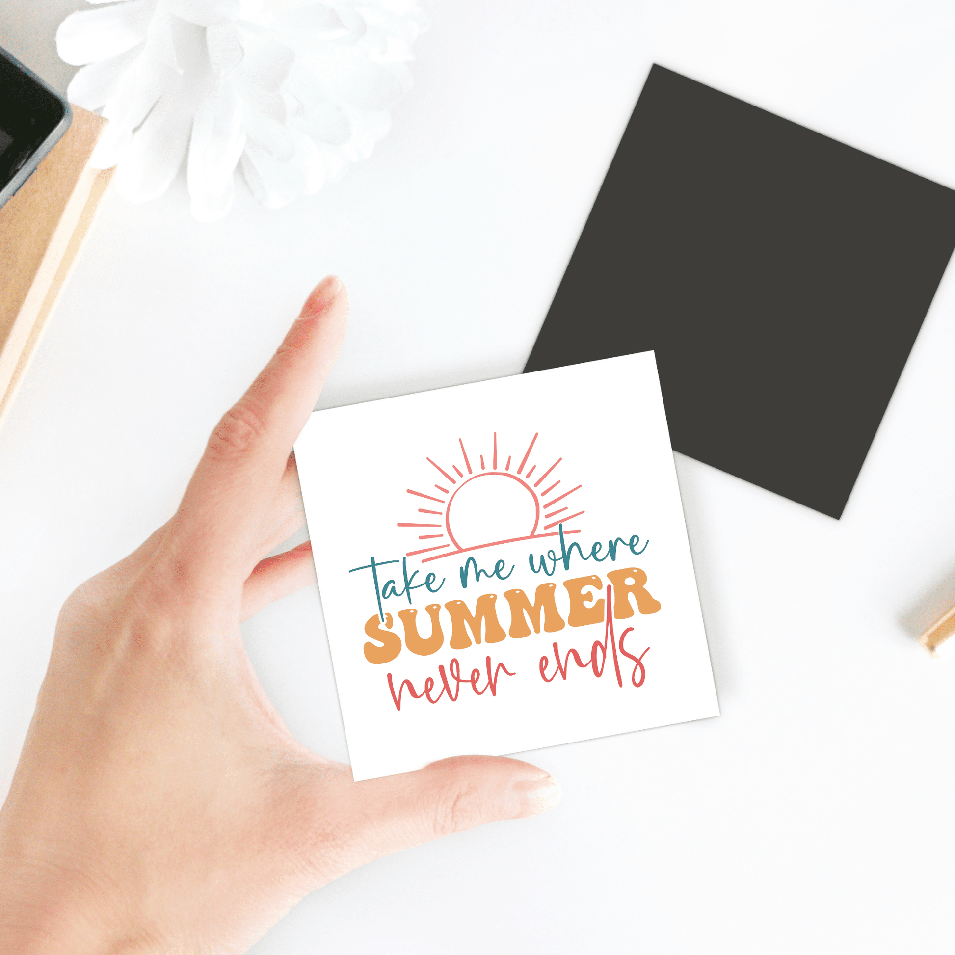 A hand holds the "Take Me Where Summer Never Ends Magnet" by Mirabilia Boutique, featuring a sun illustration—ideal for summer or travel-themed fridge decor. Nearby are a black square, white flower, notebook, and pencil on a white surface.