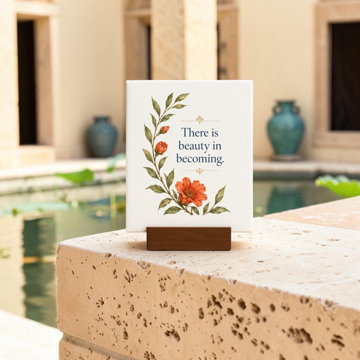 The “There Is Beauty in Becoming” Mini Canvas Sign by Mirabilia Boutique, featuring floral artwork and an inspiring quote, is displayed on a stone ledge beside a pond, with potted plants and a beige building visible in the background.
