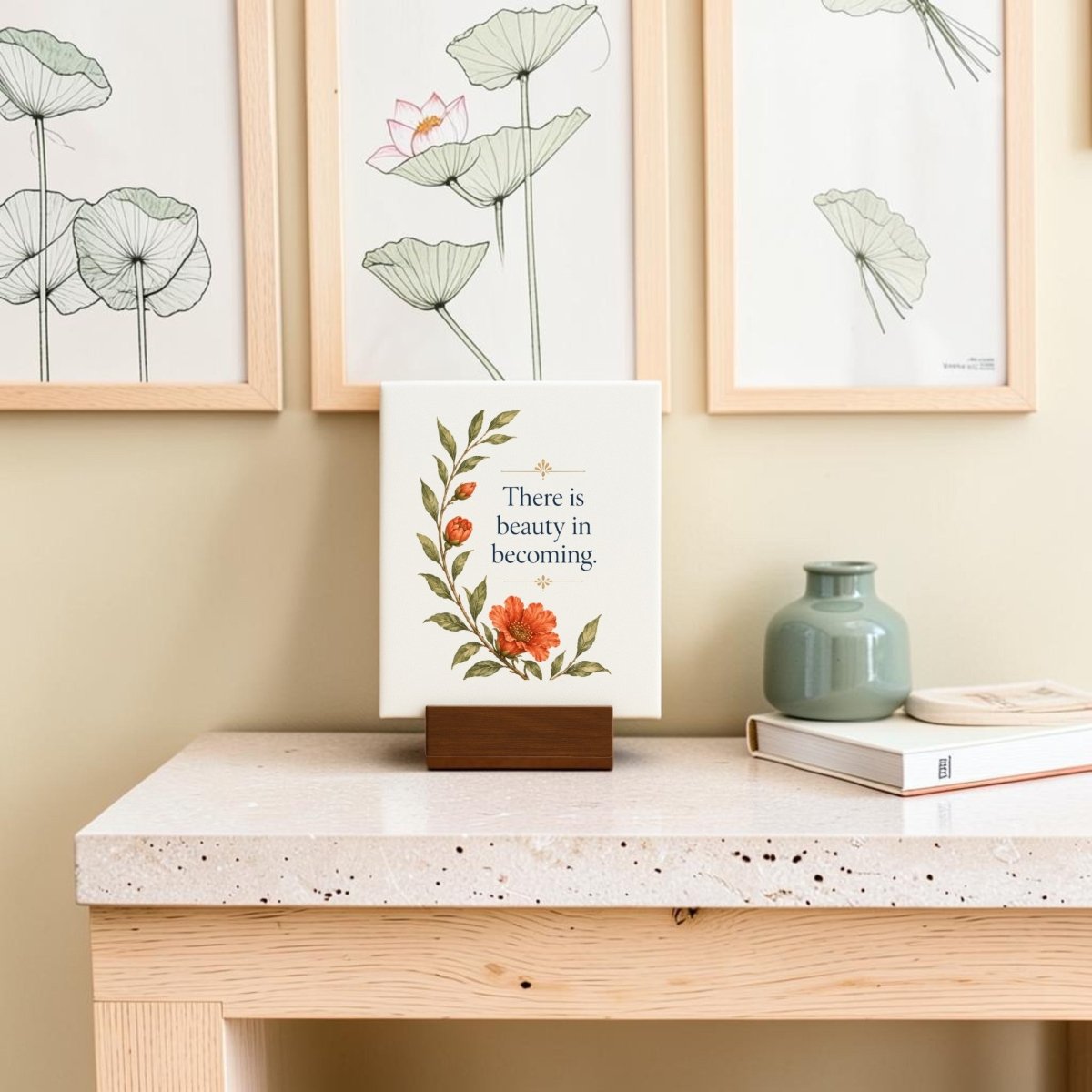 The There Is Beauty in Becoming Mini Canvas Sign by Mirabilia Boutique, featuring floral artwork and an inspiring message, stands on a wooden holder atop a light table beside a vase and a book. Framed botanical art decorates the wall behind, completing the uplifting scene.
