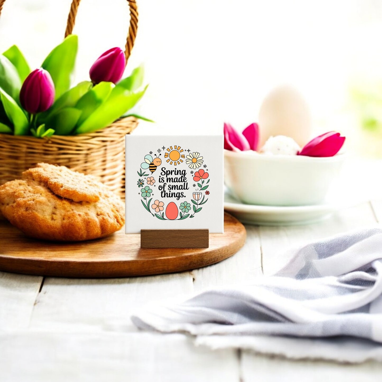 The Mirabilia Boutique "Spring Is Made of Small Things" Mini Canvas Sign sits on a table with cookies, eggs, pink tulips, and a striped cloth—a charming touch for your spring décor in natural light.