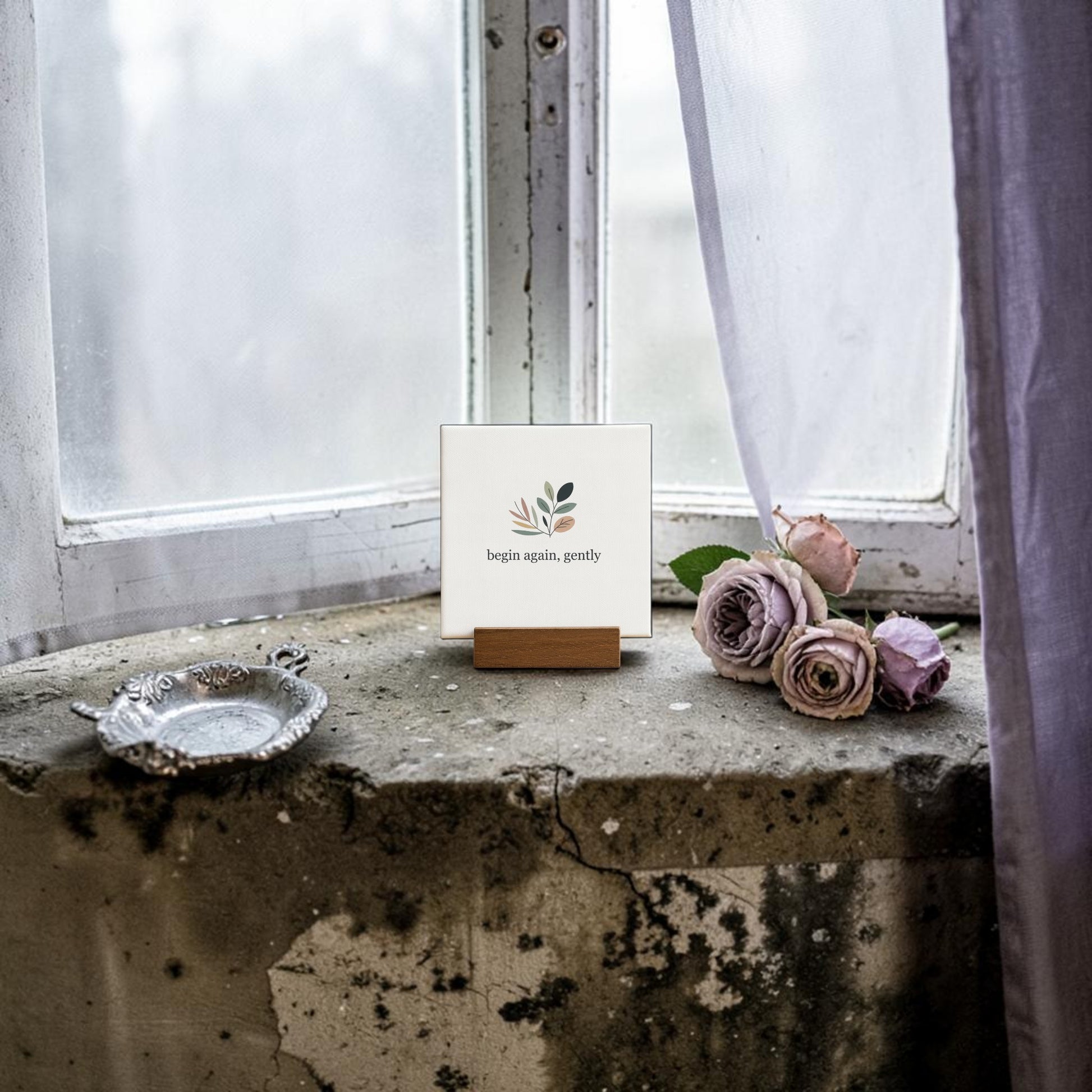 The Begin Again Gently Mini Sign with Wooden Stand by Mirabilia Boutique rests on a window ledge, ideal as botanical desk decor or a charming cottagecore gift. Soft light glows through sheer curtains beside mauve roses and a silver tray.
