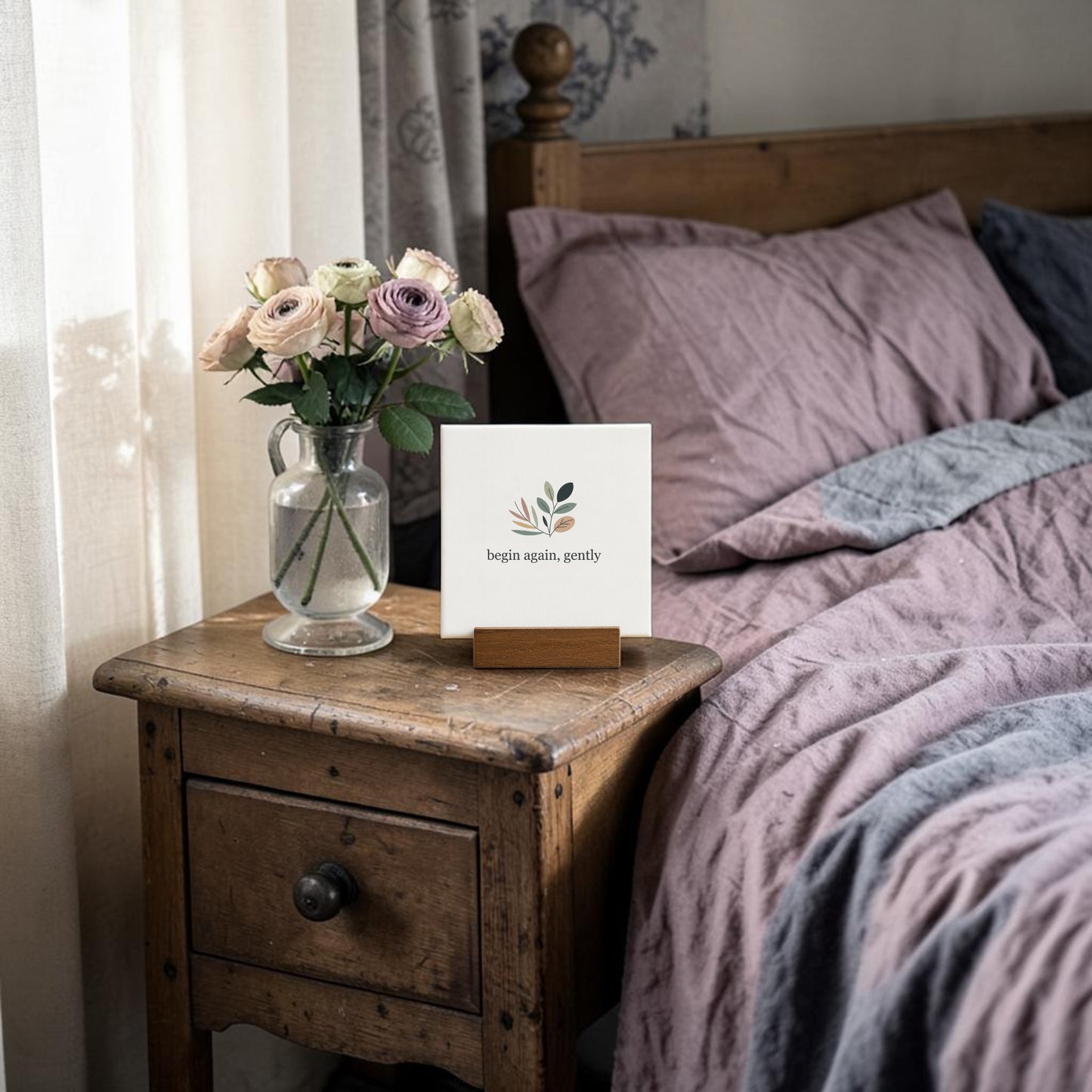 The Mirabilia Boutique Begin Again Gently Mini Sign with Wooden Stand sits on a rustic nightstand beside a glass vase of pastel flowers, perfect as cottagecore decor or a botanical gift near a bed with mauve and gray bedding by sheer-curtained windows.