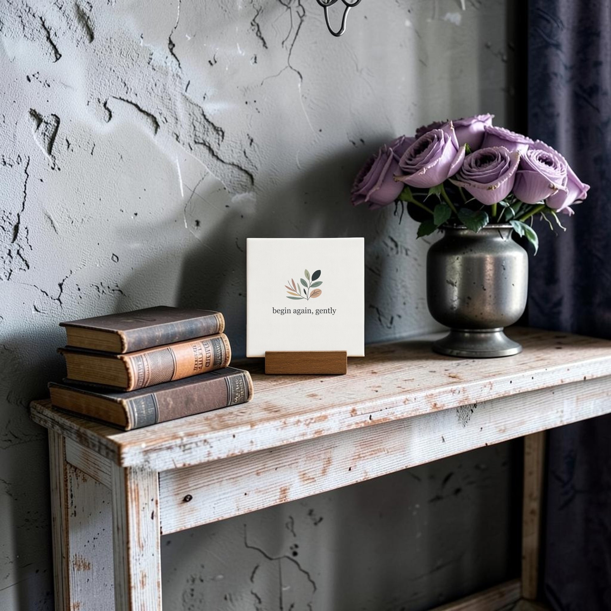 A rustic wooden table displays Mirabilia Boutique’s Begin Again Gently Mini Sign with Wooden Stand, surrounded by vintage books and a metal vase of purple roses—an enchanting cottagecore gift set against a textured gray wall.