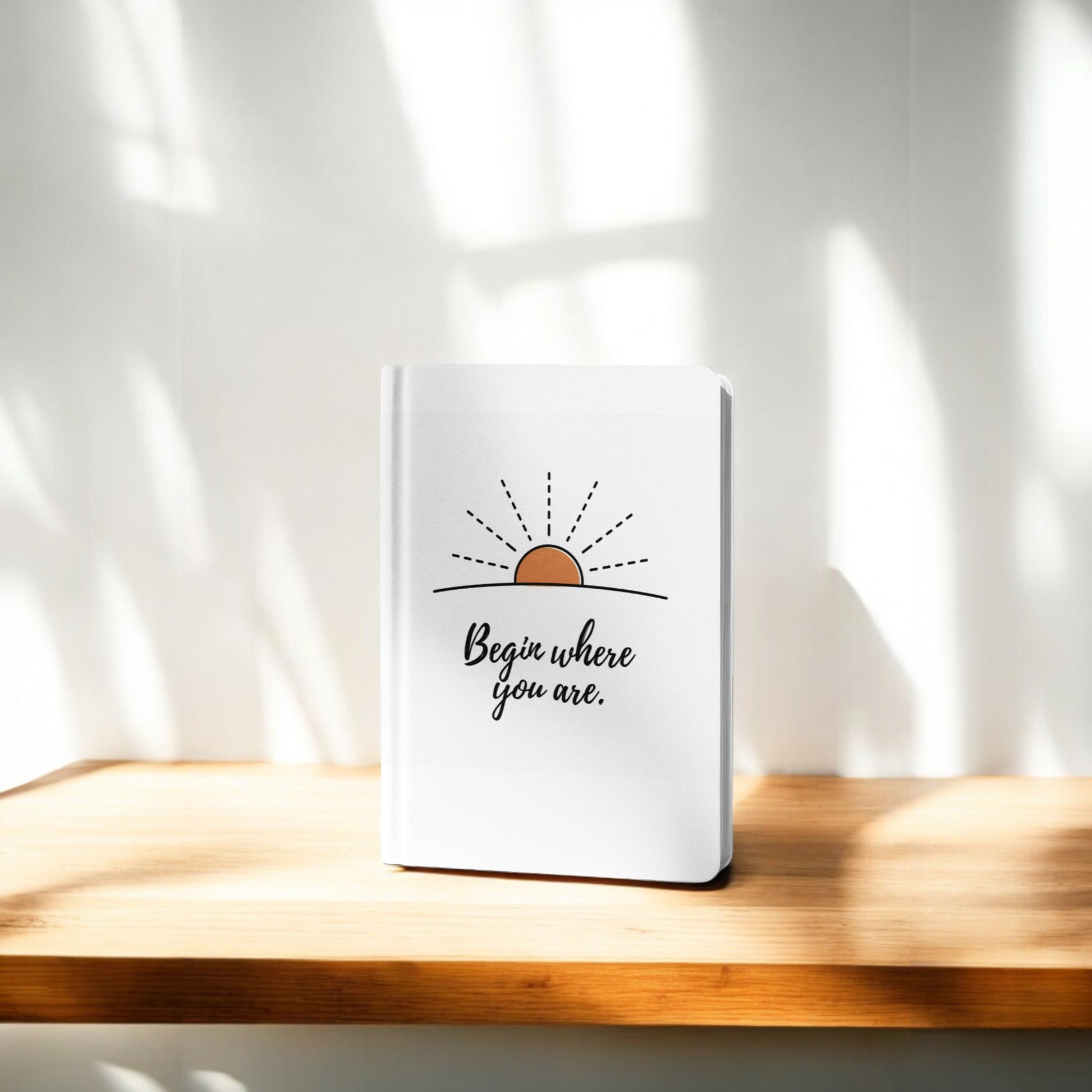 The Mirabilia Boutique "Begin Where You Are Journal – Sunrise Quote Notebook" rests on a sunlit wooden shelf, its cover adorned with a sunrise illustration and inspiring words—ideal for daily writing motivation.