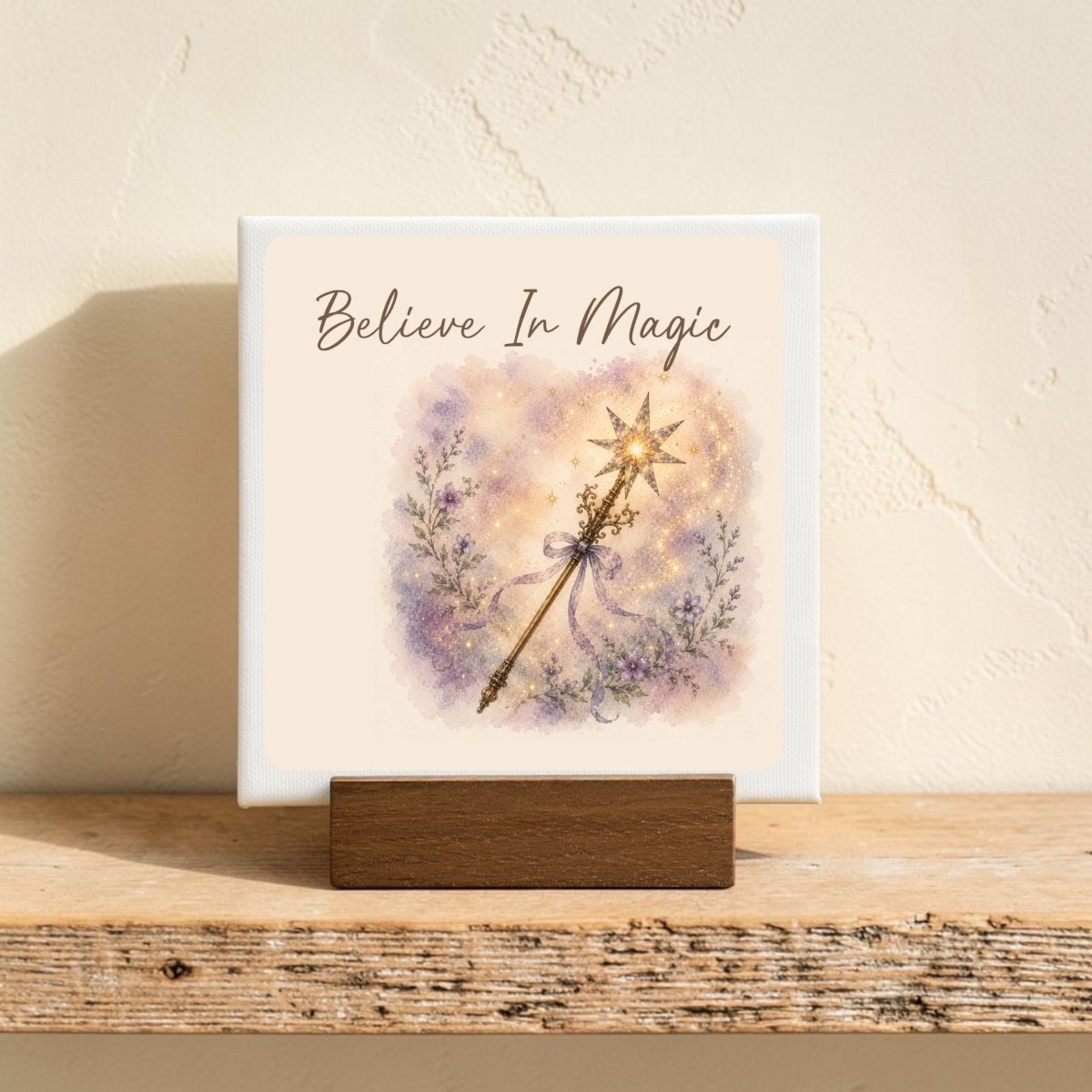 The "Believe In Magic Mini Canvas Sign" by Mirabilia Boutique features the uplifting phrase “Believe In Magic” above a sparkling illustrated fairy wand and delicate purple flowers, evoking the enchanting charm of watercolor floral decor. This small canvas sign is displayed on a wooden stand against a softly textured light wall.
