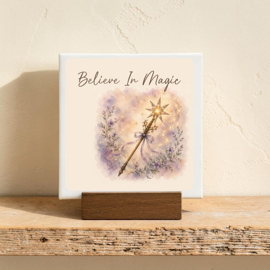 The "Believe In Magic Mini Canvas Sign" by Mirabilia Boutique features the uplifting phrase “Believe In Magic” above a sparkling illustrated fairy wand and delicate purple flowers, evoking the enchanting charm of watercolor floral decor. This small canvas sign is displayed on a wooden stand against a softly textured light wall.