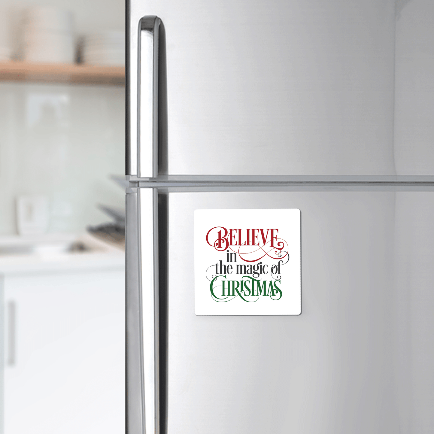 The Believe in the Magic of Christmas Magnet by Mirabilia Boutique features festive red and green lettering—perfect for holiday gifting or as a stocking stuffer. Blurred kitchen shelves and utensils appear in the background.